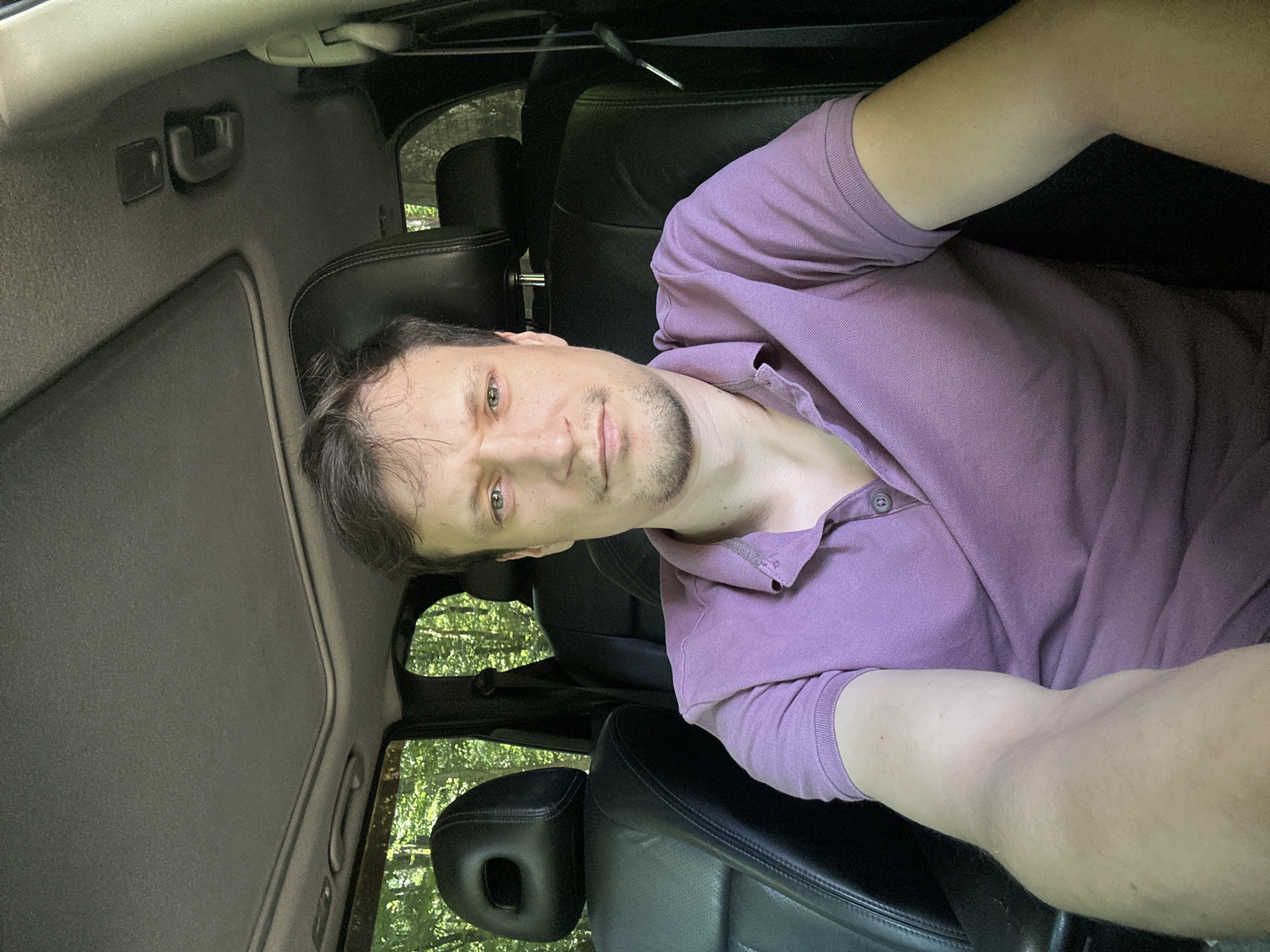A man with light brown hair and blue eyes is sitting in the driver's seat of a car. He is wearing a purple polo shirt and looking directly at the camera. The interior of the car appears to be made of black leather. Outside the car, a forest with green trees is visible through the windows and sunroof. The car is parked in Shatava, Ukraine.