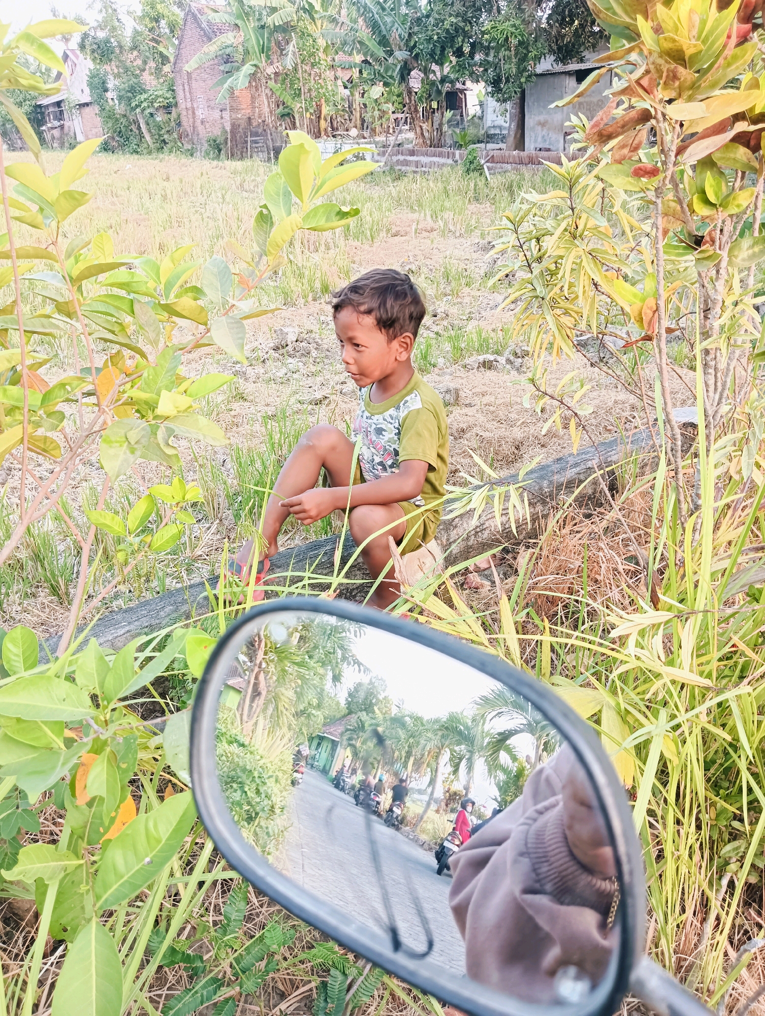 A young boy is squatting on a log in a rural outdoor setting in Sentul, Indonesia. The boy is wearing a green t-shirt and shorts. He is looking to his left. In the foreground, there is a motorcycle mirror reflecting a road with several motorcycles and people. The background shows a dry field, trees, and a building. The image has a warm, natural light.