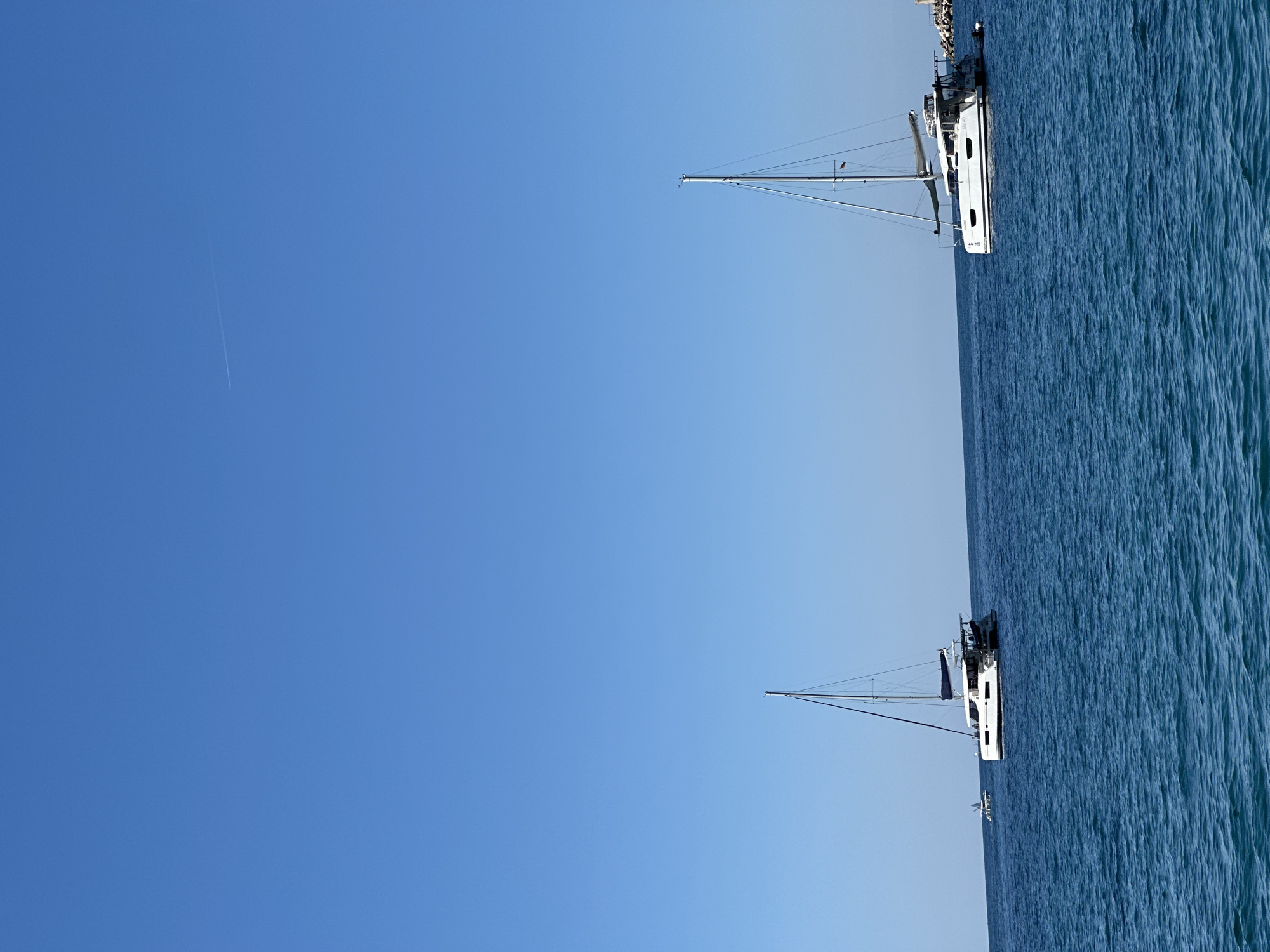The image shows two sailboats at sea under a clear blue sky. One sailboat is positioned in the upper right corner of the image, while the other is in the lower right corner. The water appears wavy. There is a contrail visible in the upper left quadrant of the sky. The scene is likely located off the coast of Budva, Montenegro.