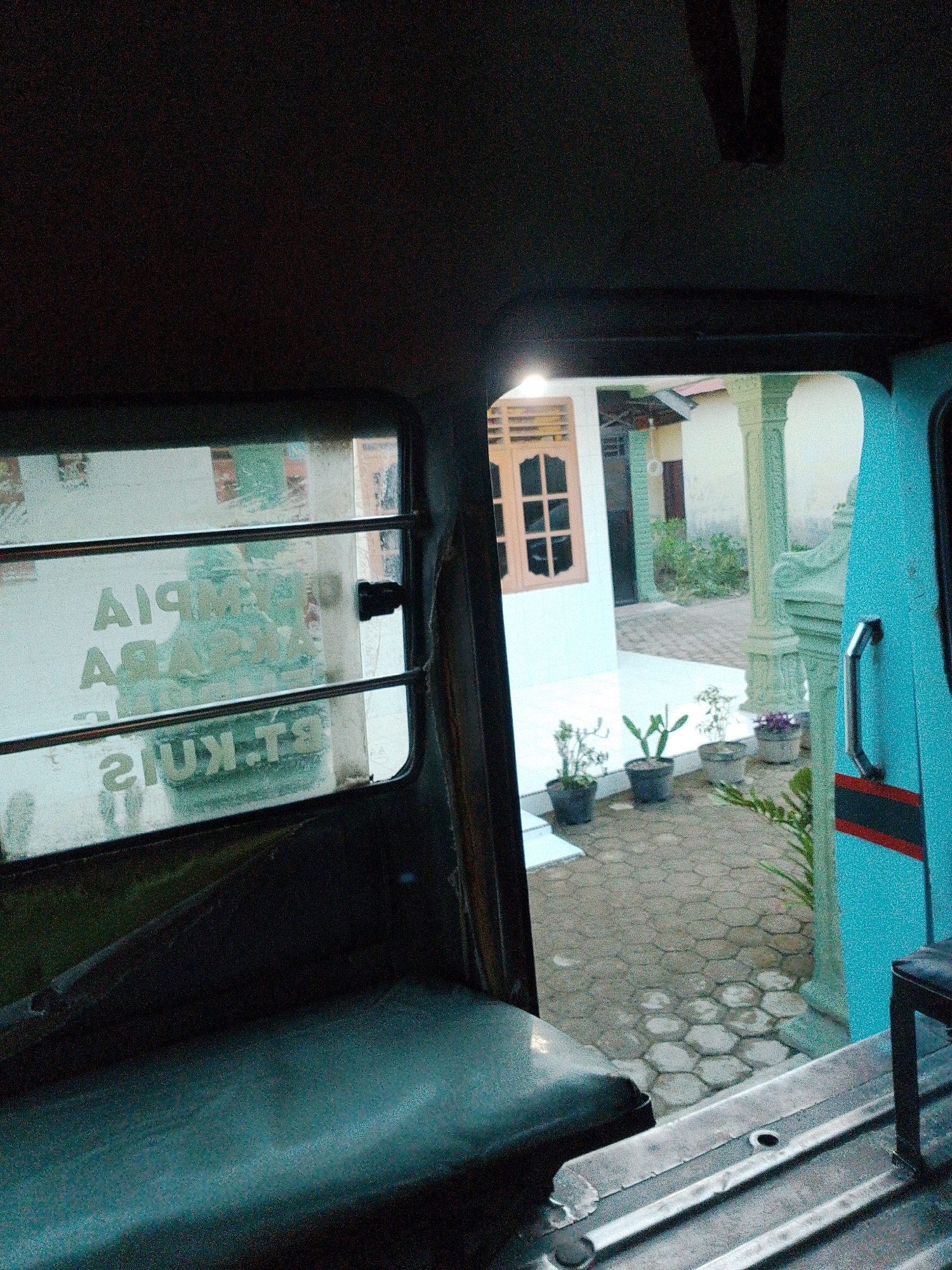 The image is taken from inside a vehicle, looking out through a window at a building and an outdoor area. The window has horizontal bars across it, and some text is visible on the glass, which appears to be in Indonesian. Outside the window, there is a building with a light-colored exterior and a wooden-framed window. In front of the building, there is a paved area with hexagonal tiles, and several potted plants are arranged on the ground. Some architectural elements, possibly pillars, are also visible. The overall impression is of a scene in a residential or commercial area in Bandar Khalipah, Indonesia. The lighting suggests it might be daytime, possibly overcast or late afternoon, given the subdued light. There are no people clearly visible in the image. The interior of the vehicle appears functional, with a dark-colored seat visible in the foreground and metal components of the vehicle's structure. The exterior of the vehicle, where visible on the right, is painted a bright blue with a red stripe.