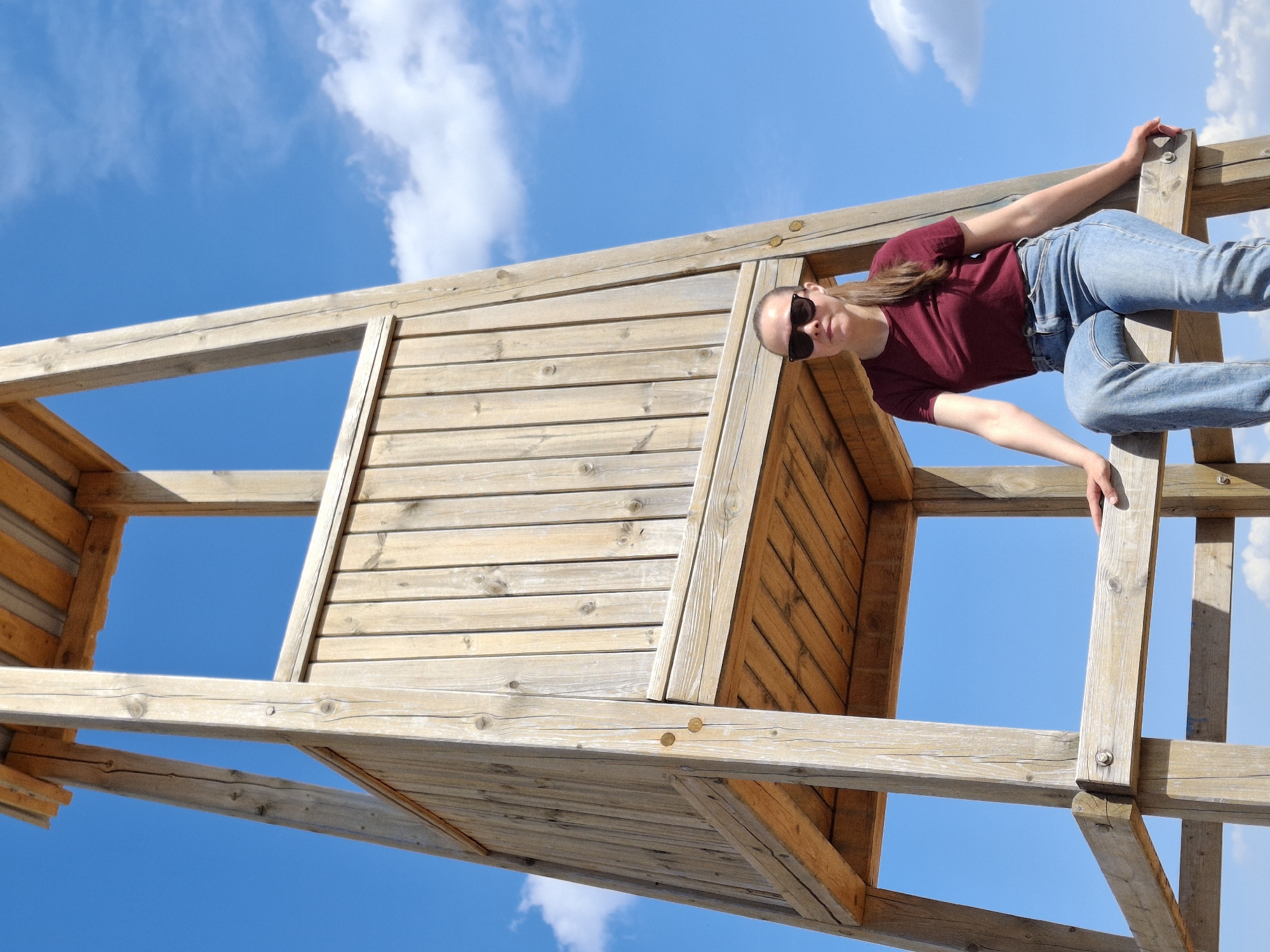 The image shows a woman climbing on a wooden play structure. The structure appears to be in a playground or park. The woman is wearing jeans, a dark red shirt, and sunglasses. She is holding onto the wooden beams of the structure. The background is a blue sky with some white clouds. The location is Chaikovsky, Russia.