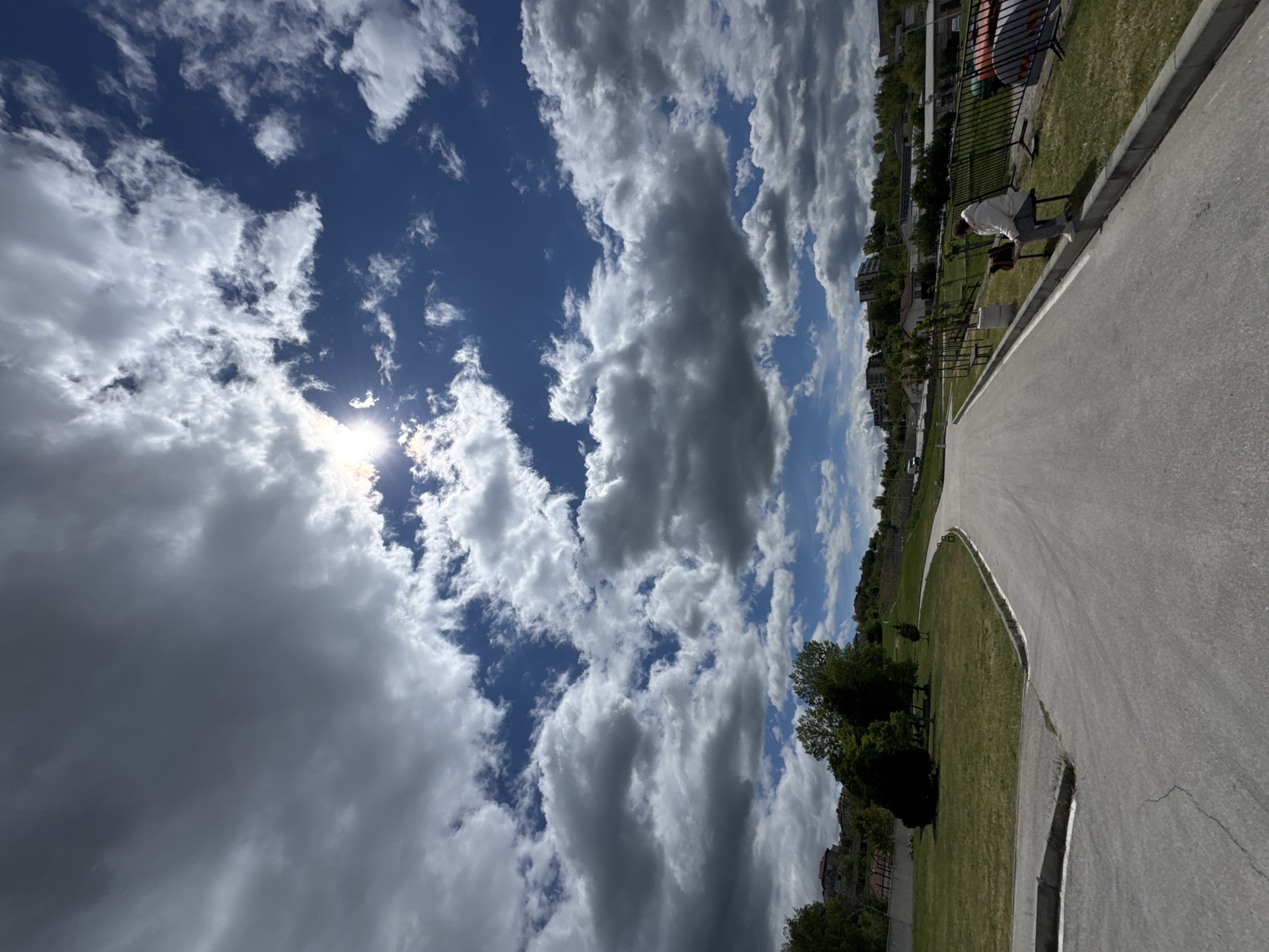 The image captures a scene in Blagoevgrad, Bulgaria, showcasing a partly cloudy sky above a city park. The sun shines through a break in the clouds. A paved walkway curves through the park, bordering a grassy area with trees and foliage. Buildings are visible in the background, indicating an urban environment. A person is bent over sitting on a bench near the path. A black metal fence is behind the bench.