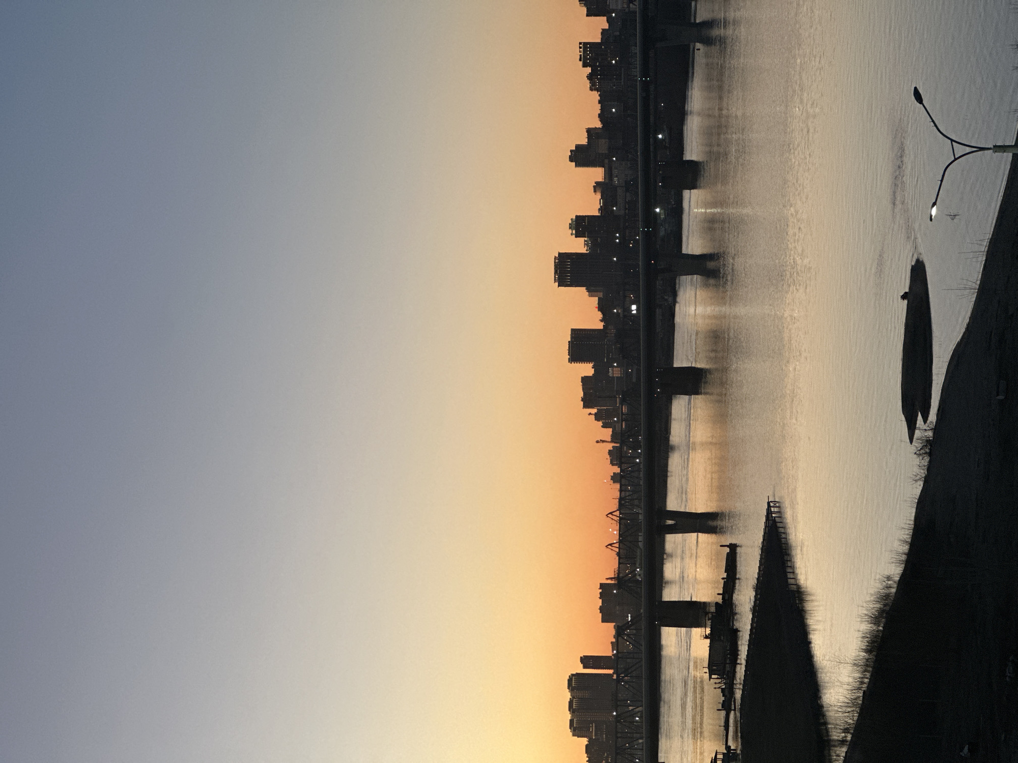 The image depicts a tranquil urban landscape in Novosibirsk, Russia, captured during either sunrise or sunset, evident by the gradient sky and silhouetted forms. The sky transitions from a soft, pale blue in the upper left portion of the frame to warmer hues of orange and yellow towards the horizon.
Across the mid-ground, a sprawling city skyline is silhouetted against the colorful sky. Numerous buildings, including high-rises and residential structures, create a jagged, dark profile. A prominent bridge, likely spanning the Ob River, stretches across the scene, its structural elements and distant lights faintly visible in silhouette.
The lower half of the image is dominated by the wide, reflective surface of the river. The water shimmers with the reflected light of the sky, showing subtle ripples and patterns. On the right-hand side of the foreground, a dark, indistinct riverbank rises, featuring sparse vegetation and a modern, illuminated street light with two heads, casting a soft glow. Further down the river in the foreground, other dark, silhouetted structures, possibly docks or remnants of infrastructure, can be seen.
No individuals or discernible text are visible in the scene, which conveys a sense of calm and atmospheric beauty at the transition between day and night.