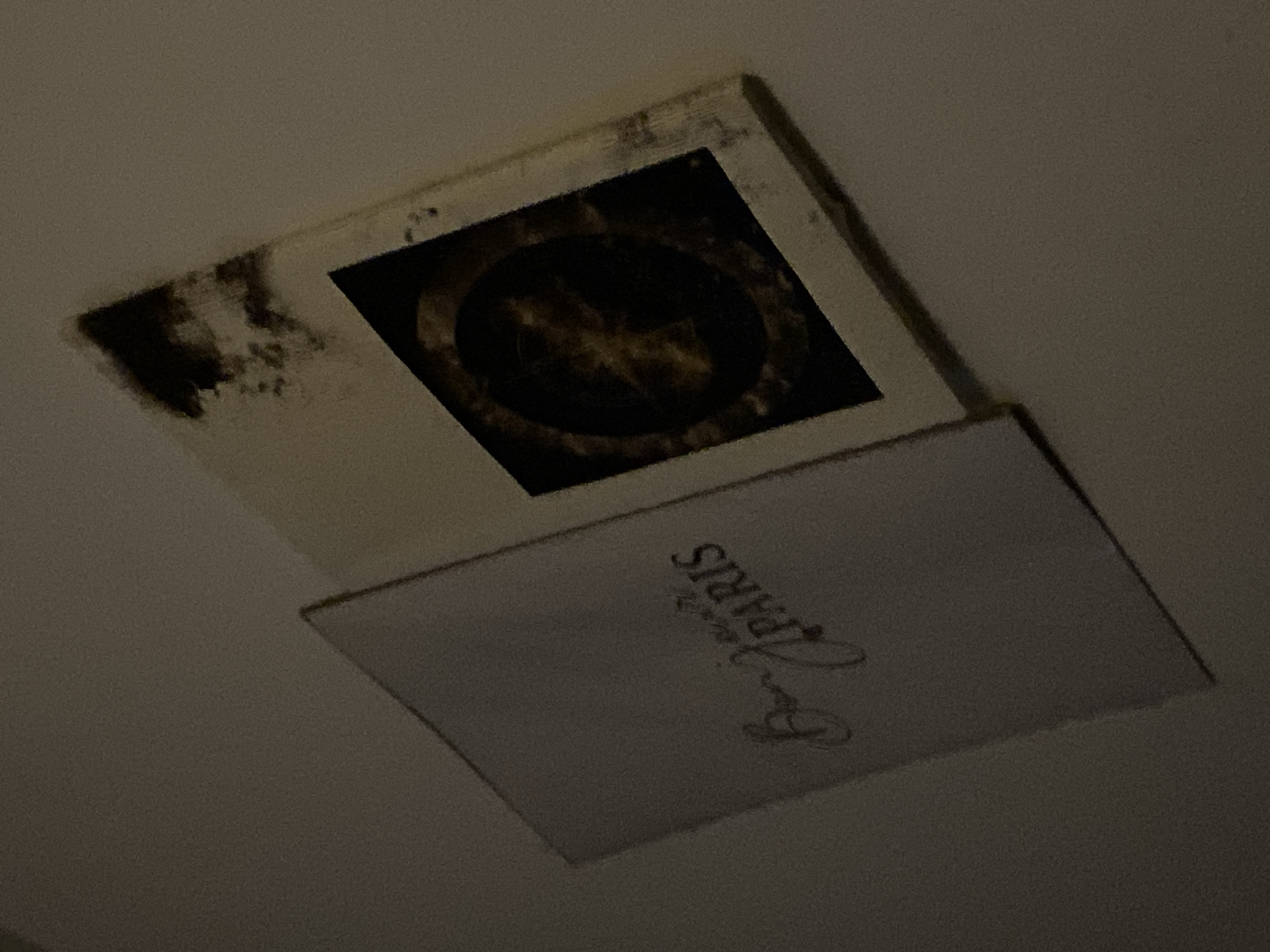 This is an overhead, eye-level, medium shot of an open box on a light-colored surface. The box appears to be made of cardboard. The left side of the box is closed, and the words "Bonjour PARIS" are inscribed on it in a script font. The right side of the box is open, revealing the inside. On the interior of the right side of the box, there is a circular graphic. The graphic has a dark background with lighter-colored, abstract shapes and patterns within it. The overall lighting of the image is dim, with some areas of shadow.