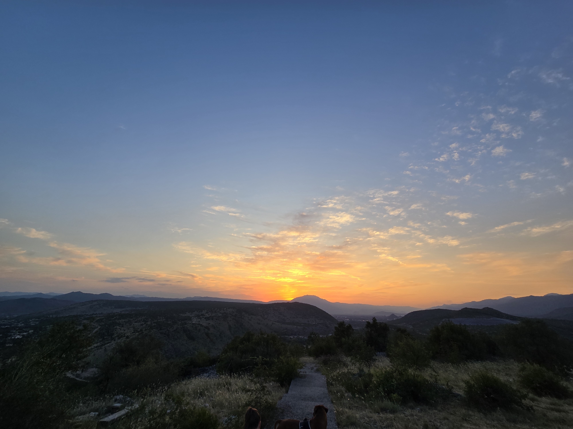 A wide-angle shot captures a panoramic view of the city of Podgorica, Montenegro at sunset. The sky transitions from a deep blue at the top to a vibrant orange and yellow near the horizon, with scattered clouds catching the warm light. Rolling hills and mountains form the landscape, with dark silhouettes of trees and vegetation in the foreground. A concrete pathway leads towards the distant mountains, and two figures, one person and one dog, are visible from behind, facing the sunset. The scene conveys a sense of tranquility and natural beauty as day transitions into night.