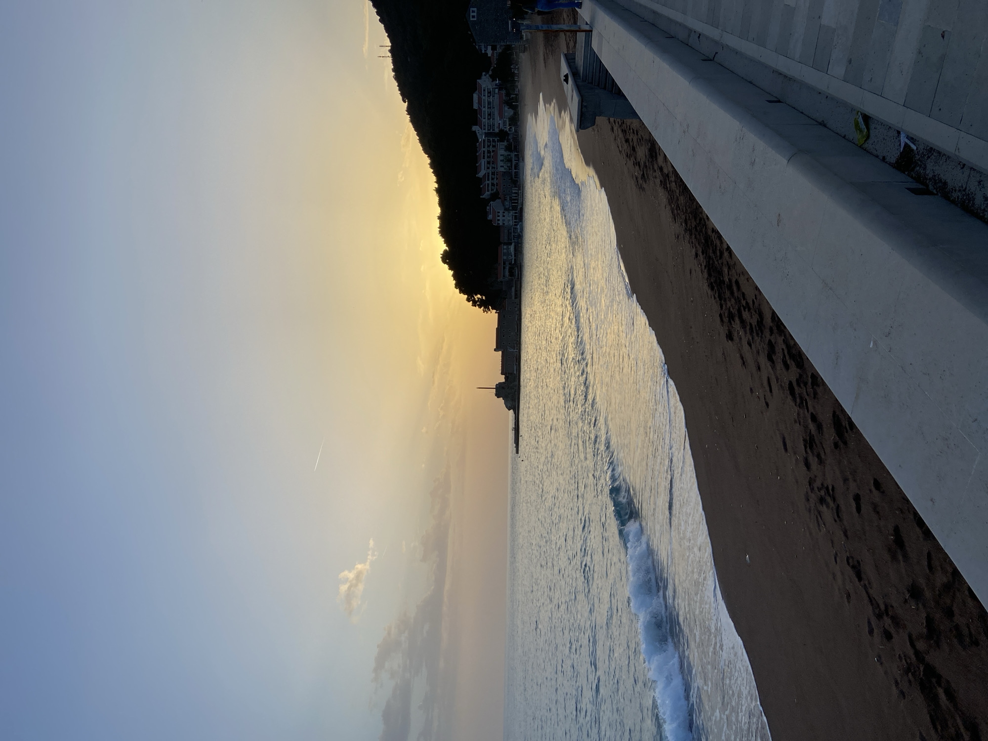 The image shows a beach scene at dawn or dusk in Petrovac centar, Montenegro. A dark, possibly volcanic sand beach is lined with waves. A concrete barrier runs along the edge of the beach. Beyond the beach, there is a hillside covered with buildings, dark green trees, and perhaps some dark buildings. The sky transitions from blue to golden yellow, suggesting sunrise or sunset. There are light clouds in the sky and what appears to be a contrail of an airplane. A tall structure that might be a communication tower or a part of a building is visible on the hillside. There are no people visible in the image.