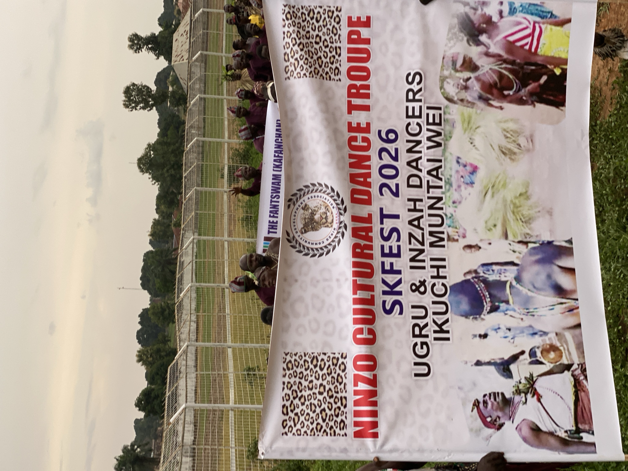 The image captures a banner displaying the text "NINZO CULTURAL DANCE TROUPE SKFEST 2026 UGRU & INZAH DANCERS IKUCHI MUNTAI WEI." The banner also includes images of people performing cultural dances.
A group of people stands behind the banner, some wearing traditional headwear. A chain-link fence stretches in the background. Trees and a cloudy sky are visible above the fence. The banner is placed in the city of Kafanchan, Nigeria. The text on the banner also contains the words "THE FANTSWAM (KAFANCHAN)."