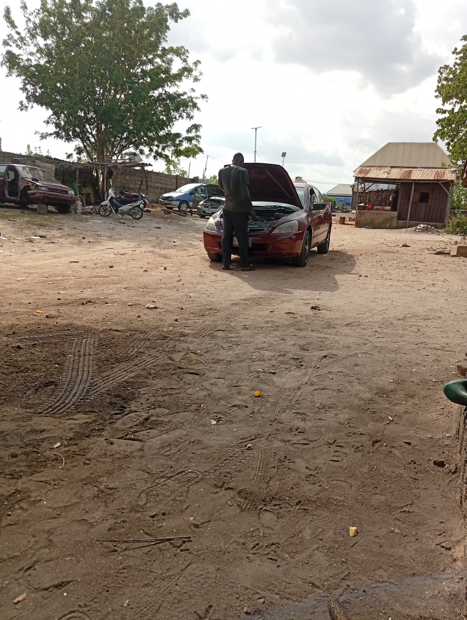 A man in dark clothing is examining the engine of a red car with its hood open in Adankolo, Nigeria. The scene appears to be a dusty outdoor workspace or auto repair area. Other vehicles are visible in the background, including a blue car, a motorcycle, and a red vehicle frame. A wooden structure stands to the right of the red car. There are tire tracks and footprints in the foreground dirt. The sky is overcast.