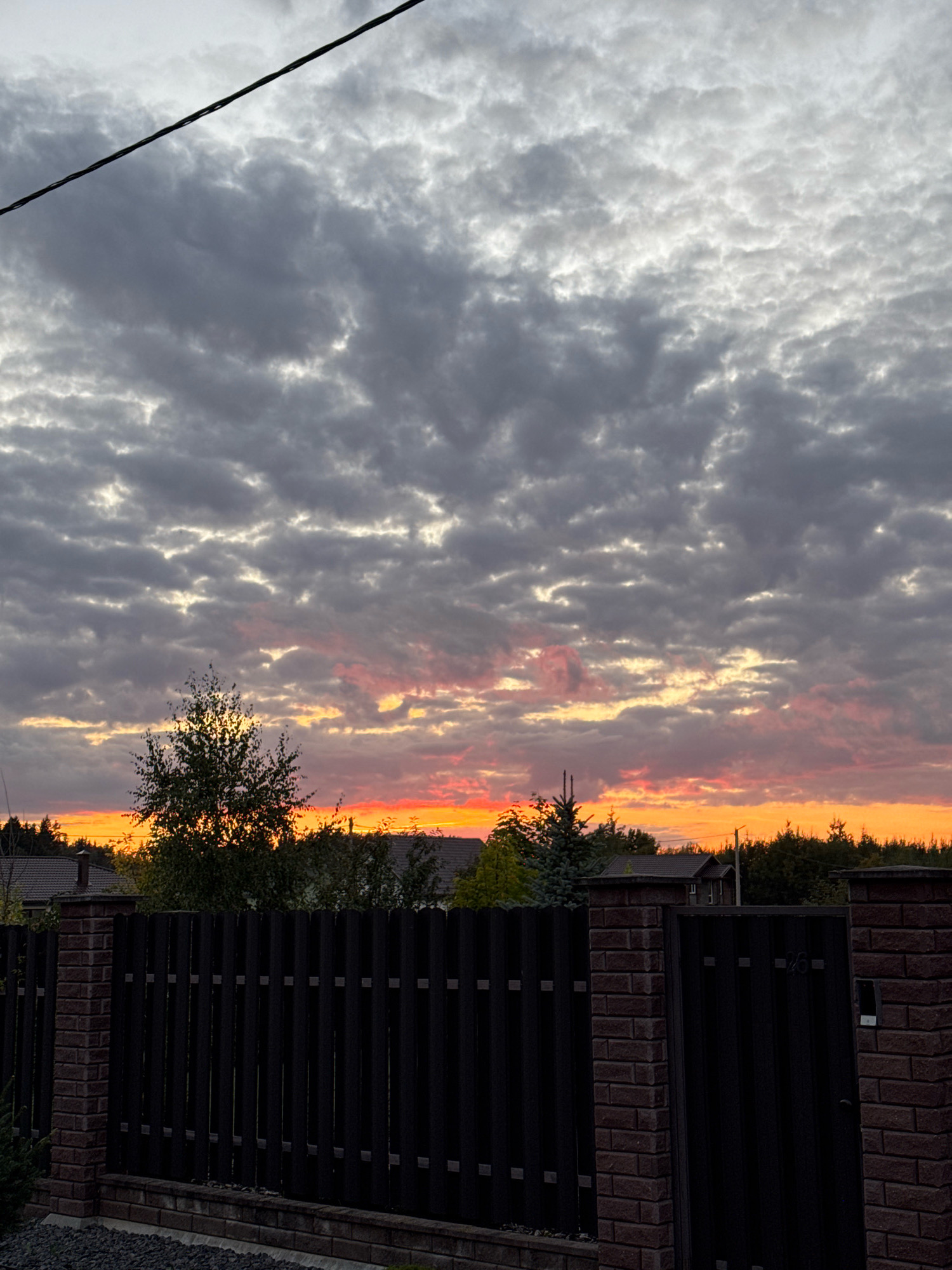 The image captures a twilight sky over a suburban area in Бараўлянскі сельскі Савет, Belarus. The sky is filled with a dramatic display of clouds, with dark gray formations interspersed with streaks of pink and orange from the setting sun. A single power line traverses the upper portion of the frame. In the foreground, a dark, vertical slatted fence with brick pillars marks the boundary of a property. Beyond the fence, silhouetted trees and residential rooftops are visible against the vibrant sky. The overall impression is one of a tranquil evening scene in a rural or semi-rural setting.