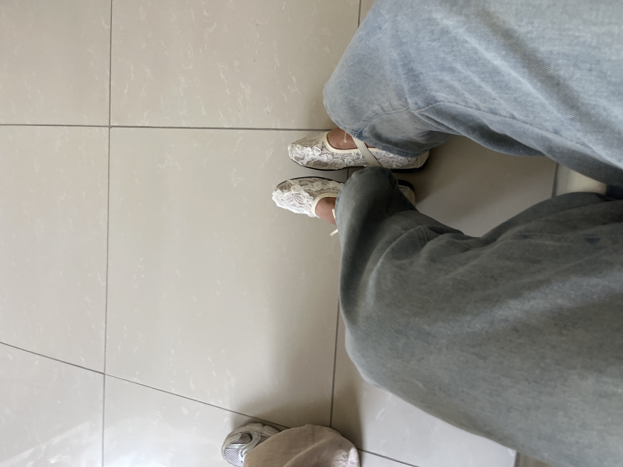The image shows a close-up view of a person's legs and feet, seated on a tiled floor. The person is wearing light-wash denim jeans and white lace ballet flats with a strap across the ankle. The floor is composed of large, square, beige tiles with subtle grey veining. The lighting suggests an indoor environment. There is a hint of another shoe and fabric on the left side of the frame, indicating the presence of at least two individuals or a person with multiple items. The scene appears to be a neutral, possibly public space, given the tiling and the casual attire. The image does not contain any elements that can be used to infer a specific location within Batken City, Kyrgyzstan.