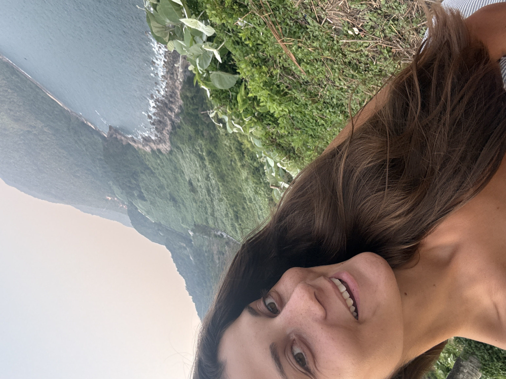 The image is a selfie of a woman with brown hair, fair skin, and freckles on her face, smiling towards the camera. She appears to be standing on a cliff overlooking the ocean. Behind her, there is a coastline with rocky cliffs, lush green vegetation covering the hillsides, and the sea. The weather appears to be overcast with a grey sky. Based on the distinctive coastal features, the location is likely in the Son Tra Peninsula in Đà Nẵng, Vietnam.