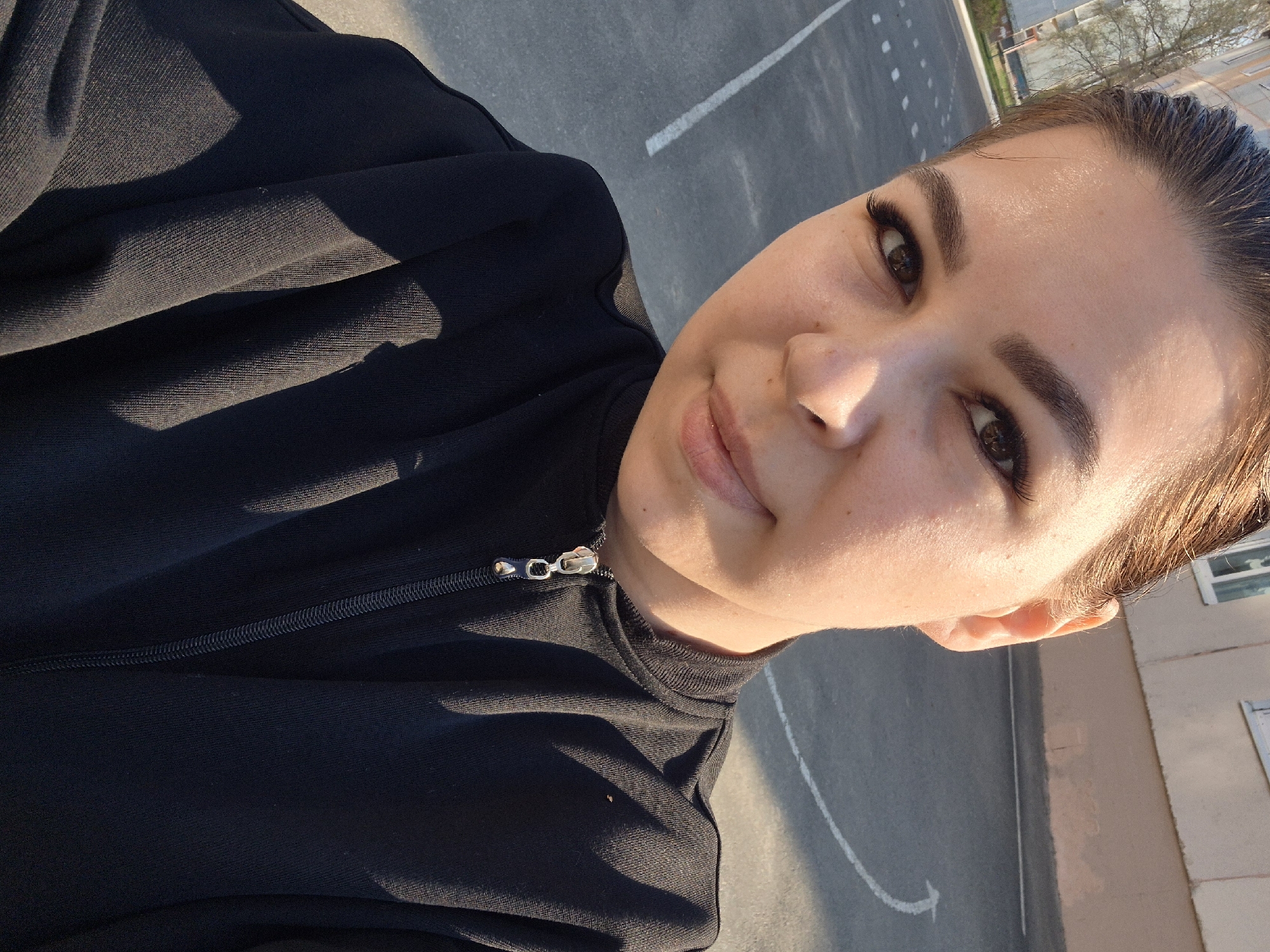 A woman takes a selfie. She wears a black zipper jacket. The background shows an asphalt surface with white lines, a building with windows, and some trees. This image seems to be taken in Tobolsk, Russia.