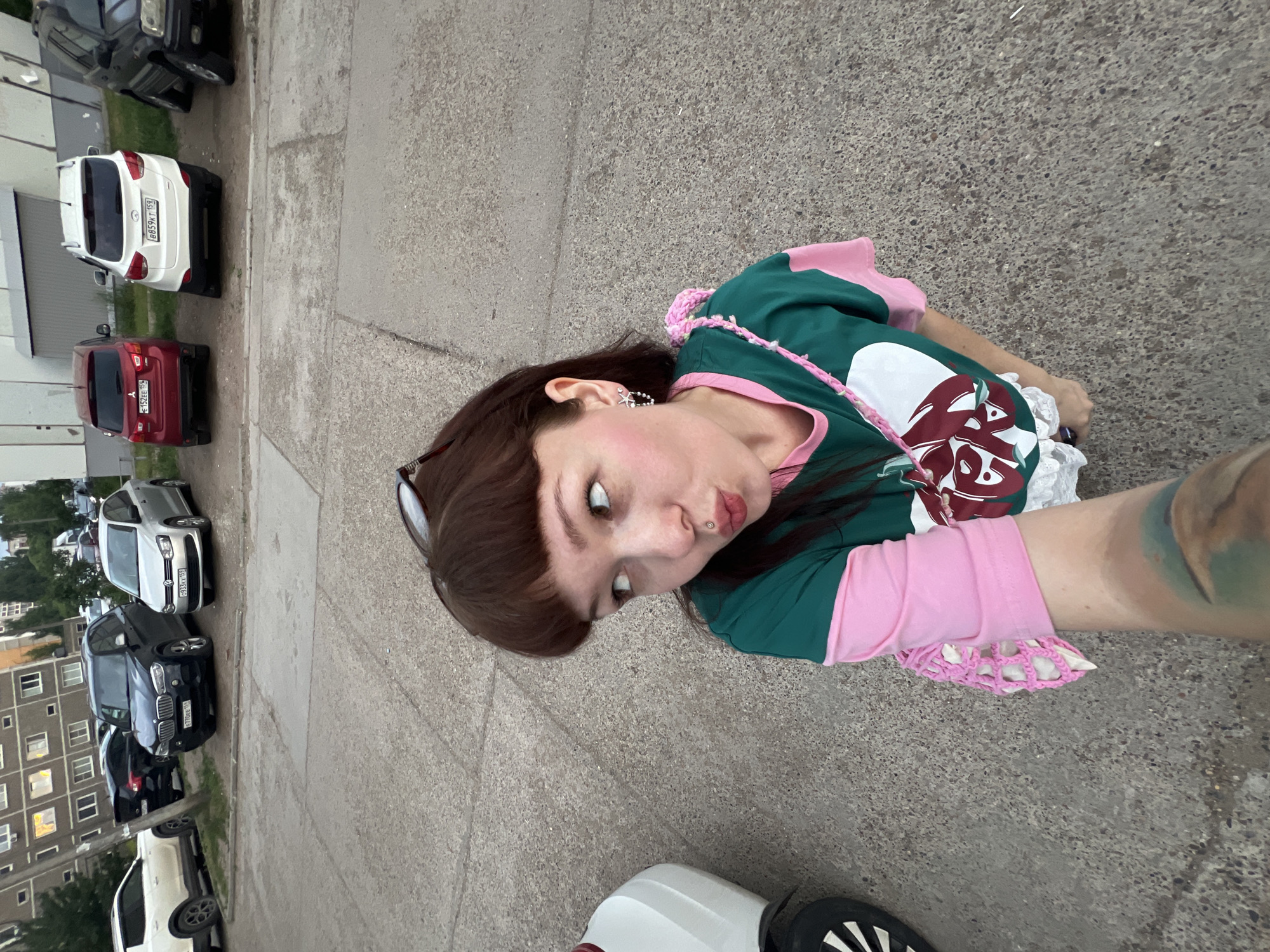 A selfie is taken by a young woman with dark brown hair, bangs, and a nose piercing, in a parking lot in the city of Chaikovsky, Russia. She is wearing a green and pink long-sleeved t-shirt and a white skirt with lace trim. A pink knitted bag is slung over her shoulder. Sunglasses are perched on top of her head. The background features several parked cars, including a silver Volkswagen Polo, a black BMW X5, a red Mitsubishi ASX, and a white Hyundai ix35. A multi-story apartment building is visible in the distance. The ground is paved with concrete.
