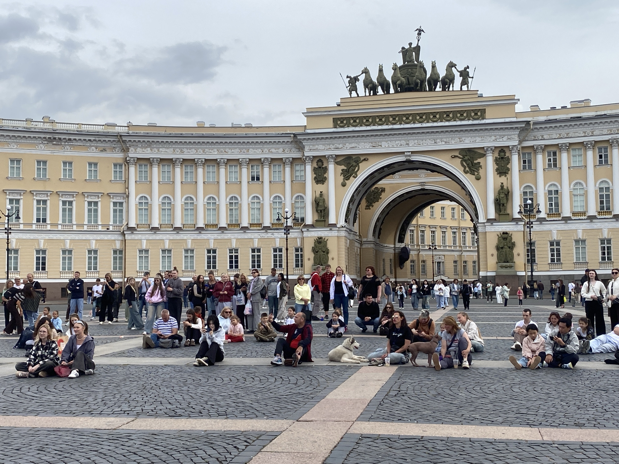 The image depicts a wide view of Palace Square in Saint Petersburg, Russia, with the General Staff Building's Triumphal Arch dominating the background. The arch is adorned with intricate sculptures, including a quadriga and various other figures. Numerous people are scattered across the cobblestone square, many of them sitting on the ground. Some individuals are engaged in conversations, while others are looking at their phones or taking pictures. Two dogs are visible among the seated individuals. The sky is overcast. The scene captures a moment of leisure and public gathering in a prominent historical location.