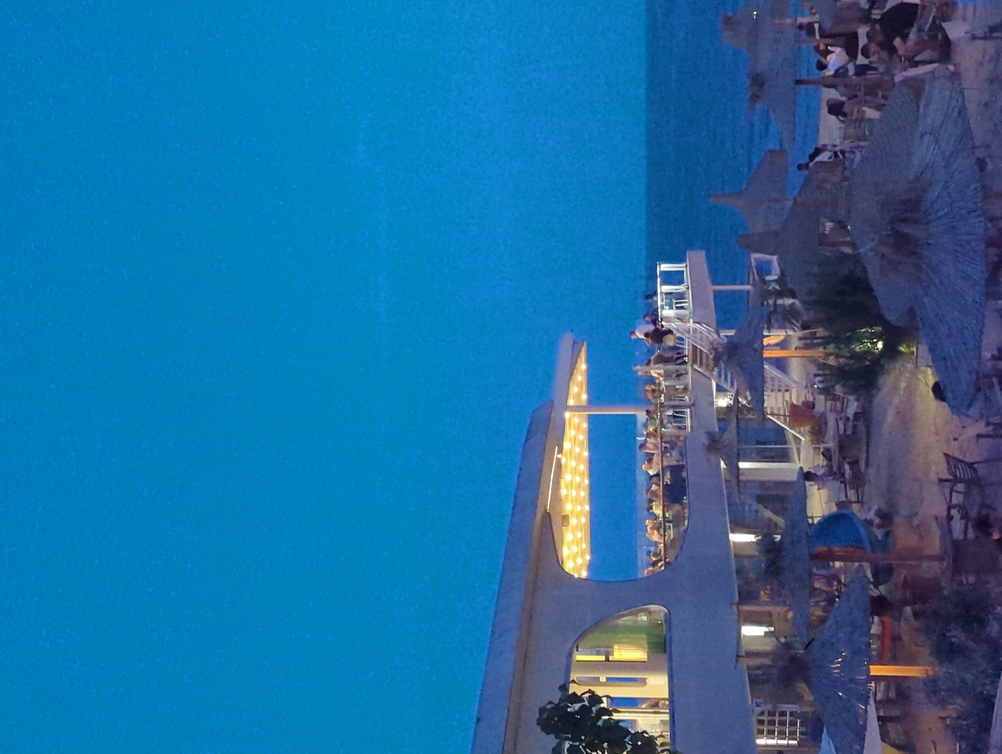 This image depicts an evening scene at a beachside restaurant or club located in Varna, Bulgaria. The sky above is a uniform deep blue, indicating either dusk or nightfall, with faint granular texture. The sea, visible on the right, matches the deep blue tone of the sky and appears calm, meeting the horizon line.
The primary structure is a multi-level building with a light-colored, modern architectural design featuring rounded edges and large, organic-shaped windows or openings. On its upper open-air balcony, numerous individuals are seated at tables under a canopy illuminated by a grid of warm yellow string lights. A white railing encloses this elevated dining area, from which a white staircase descends to lower levels. A dark green leafy plant is visible on the left side of the building's facade.
Below the main building, extending across the sandy beach, is an outdoor seating area. This area features multiple straw or thatched beach umbrellas providing shade over various tables and chairs. Several individuals are seated at these beachside tables. Artificial lighting from the building and other unseen sources softly illuminates this ground-level gathering space. A blue structure, possibly a children's slide, is partially visible among the umbrellas. People are observed in various postures, indicating social interaction and relaxation, though specific details of their activities are indistinct due to the distance and low light.