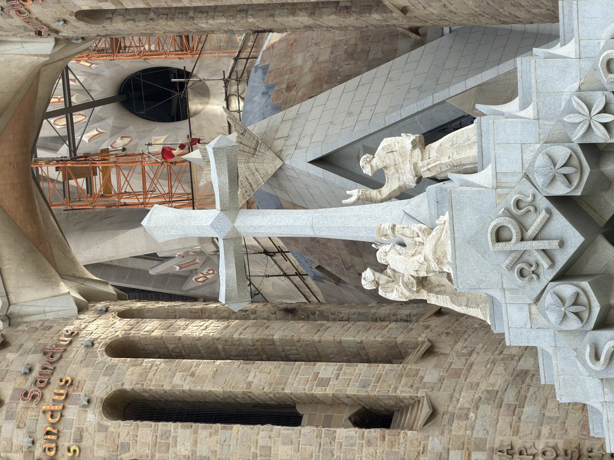 The image captures a close-up view of the Sagrada Familia basilica in Barcelona, Spain. The foreground features a large, light-gray stone cross with intricate carvings. Below the cross are stone sculptures depicting several human figures, appearing to be in prayer or contemplation. These figures are part of the basilica's facade, which is constructed from rough-hewn, tan-colored stone blocks.
The architecture is characterized by its complex and organic forms, with arched openings and textured surfaces. In the background, scaffolding and construction materials are visible, indicating ongoing work on the basilica. The overall color palette is dominated by earthy tones of stone, with hints of red and yellow in lettering on the facade, which appears to spell out "Sanctus" and possibly "Apostol". The lighting suggests a daytime scene with some shadows cast by the architectural elements.