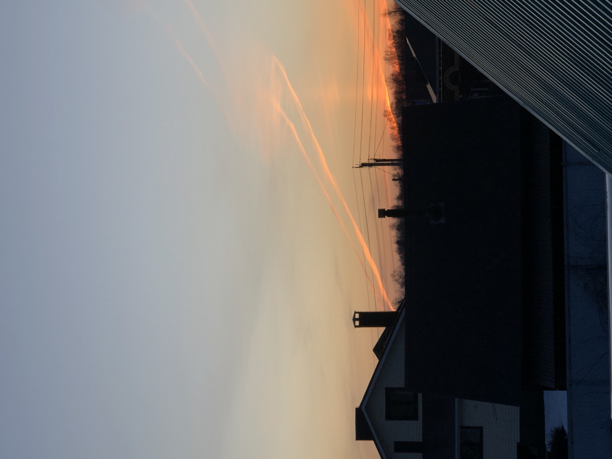 The image shows a view of the sky at sunset in Noviy, Russia. Contrails from airplanes are visible in the sky, illuminated by the setting sun. Power lines run across the frame. A residential building with a chimney and a dark-colored structure are silhouetted against the sky. A section of a corrugated metal structure is present in the foreground.