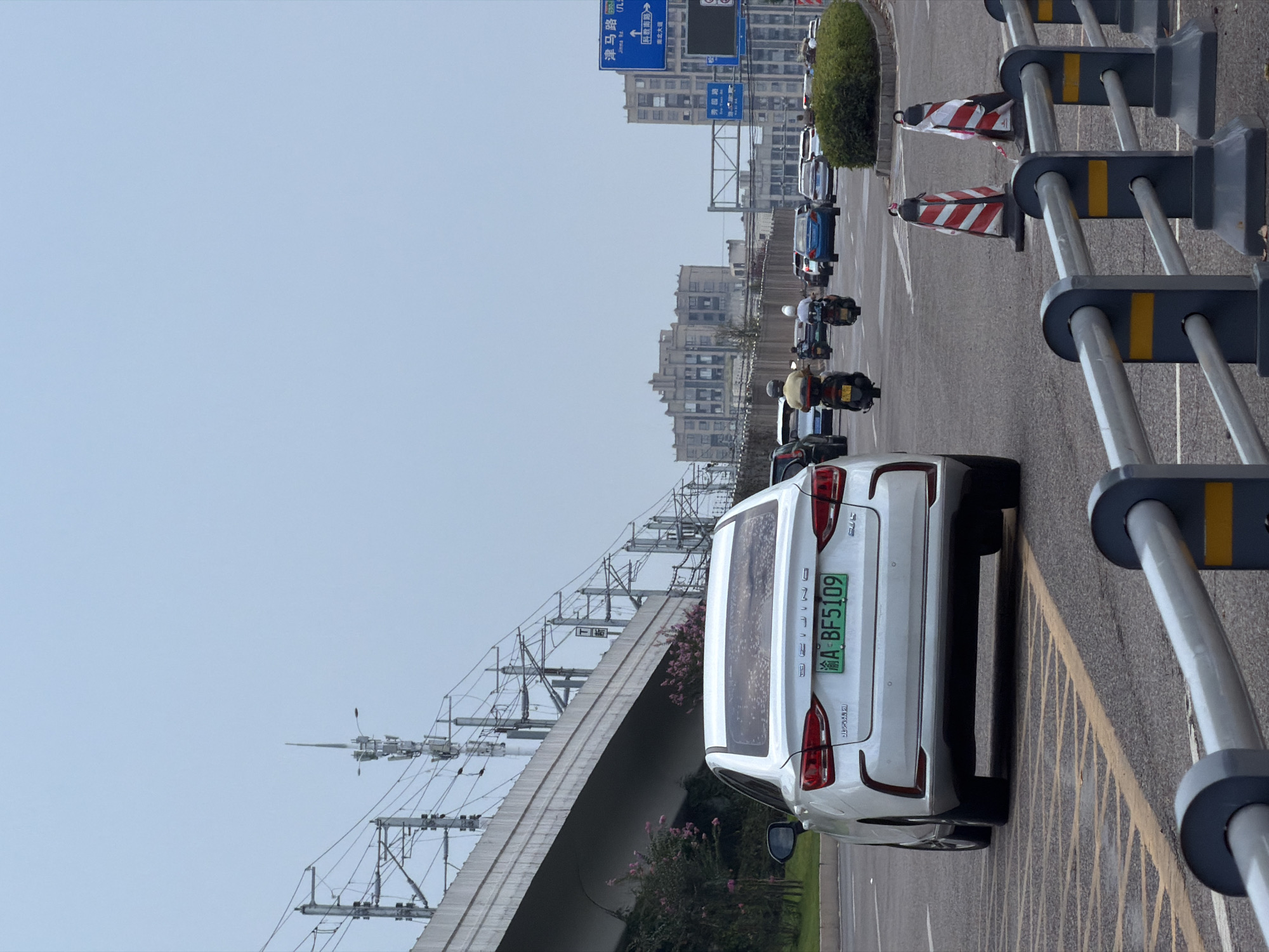 The image depicts a street scene in Jiangjin, China, under an overcast sky. In the foreground, a white sedan with the license plate "A BF5109" is prominently positioned. The car is facing away from the viewer, and its rear lights and manufacturer badging are visible. The road surface is asphalt, marked with yellow and white lines, and includes a yellow striped zone to the left of the car. A metal barrier with grey posts and silver rails, accented with yellow stripes, runs along the right side of the road.
Further down the road, several other vehicles are present, including motorcycles and cars. Two individuals on motorcycles are visible, one in yellow and one in white clothing. In the background, a large elevated railway or highway structure with overhead electrical lines dominates the left side of the frame. Numerous utility poles and antennas are also visible along this structure. Beyond the road and vehicles, several multi-story buildings are visible in the distance, with architectural styles suggesting an urban environment. Blue directional road signs are posted on the right side of the road, indicating destinations such as "Jinma Rd (九) Jiama Road" and other route information. The overall scene conveys a sense of urban traffic flow on a day with limited visibility due to cloud cover.