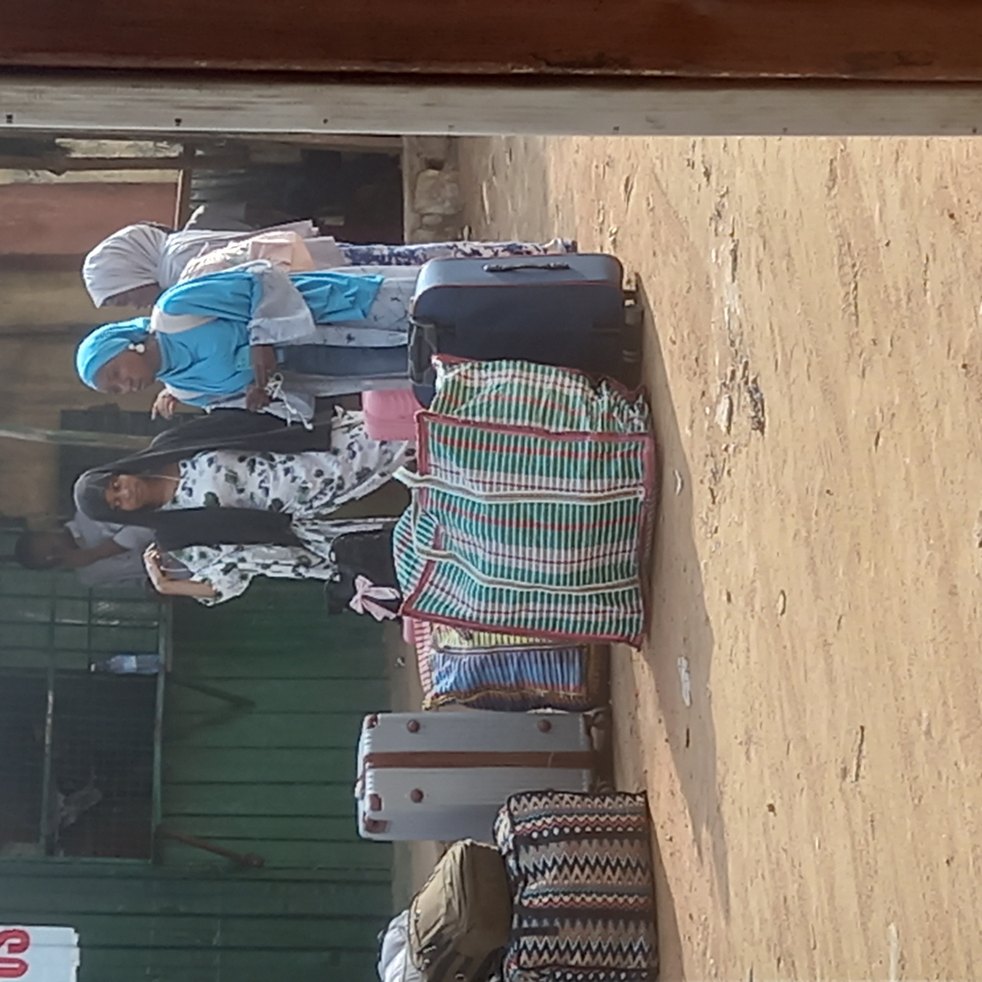 In Dadi Riba, Nigeria, several women, some wearing head coverings, stand in front of luggage. The luggage consists of suitcases and fabric bags. The surrounding environment shows a building with a green exterior and a dirt-covered ground. The scene is viewed from beneath an overhanging structure.