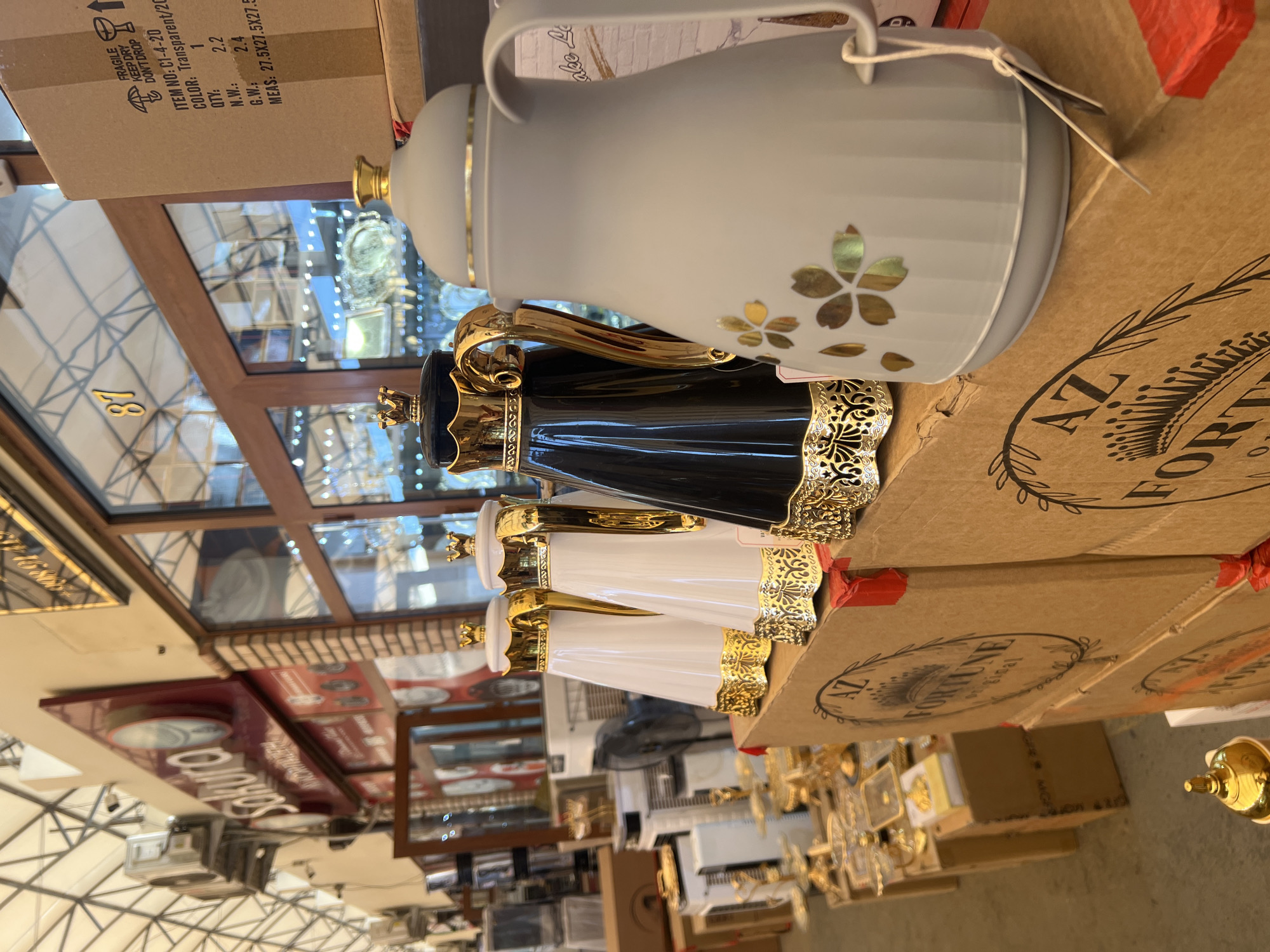 In the city of Eshanguzar, Uzbekistan, a market stall displays a variety of decorative thermoses. In the foreground, a light grey thermos with a subtle striped pattern and gold floral accents is prominent. To its left, a black thermos with ornate gold detailing and a gold crown-like lid is visible. Further left are two white thermoses, also featuring gold accents and crowns. These thermoses are placed on top of stacked cardboard boxes with the "AZ FORTUNE original" logo. In the background, a store with large windows displays more goods, and overhead, a white, tent-like structure covers the market area. Various appliances and shelves filled with items are also discernible in the background.