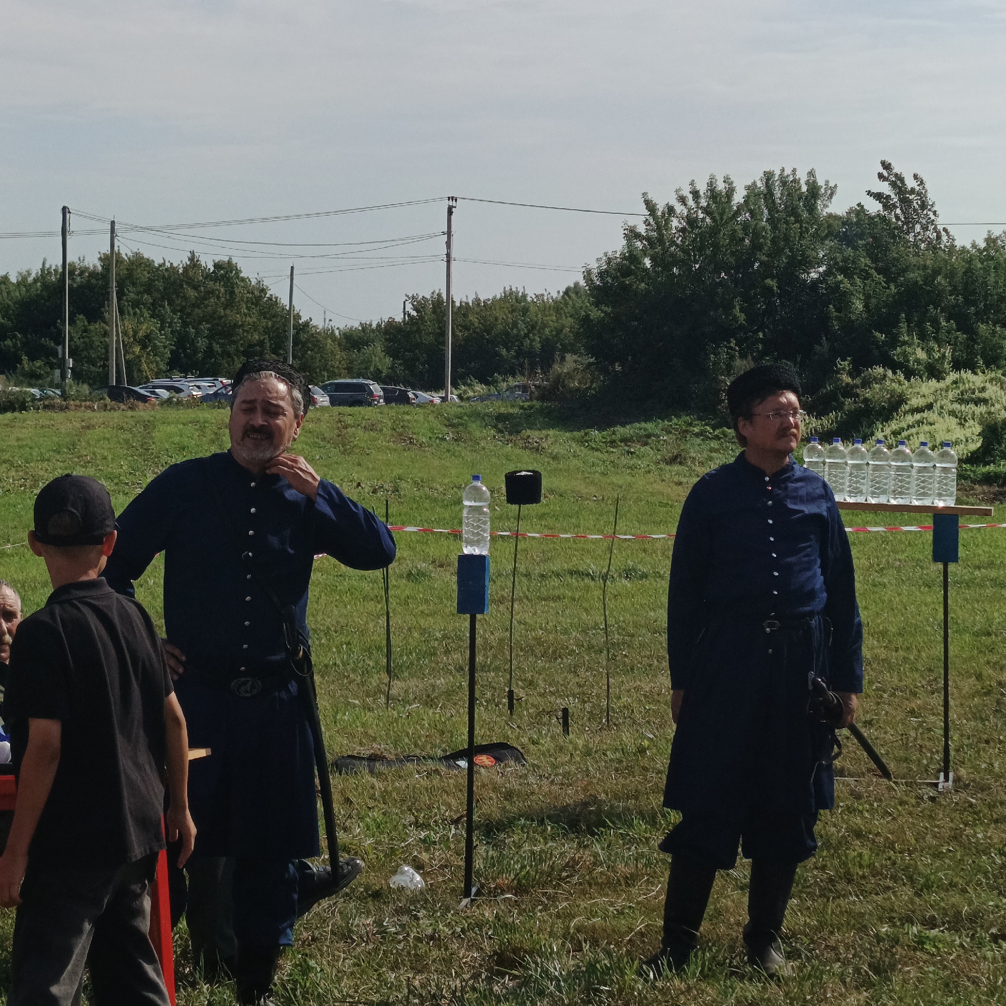 In Nikolo-Beryozovka, Russia, a group of people are gathered in a grassy field under a cloudy sky. Two men in traditional dark blue uniforms with hats are standing. One man, with a mustache and graying hair, is facing left, touching his neck and speaking. The other man, wearing glasses, is looking to the right. In the background, a row of plastic bottles is lined up on a wooden plank, presumably for a target practice event. A child in a black t-shirt and grey pants stands to the left, facing away from the camera. The environment is a rural outdoor setting with trees and parked cars visible in the distance, suggesting a community gathering or event.