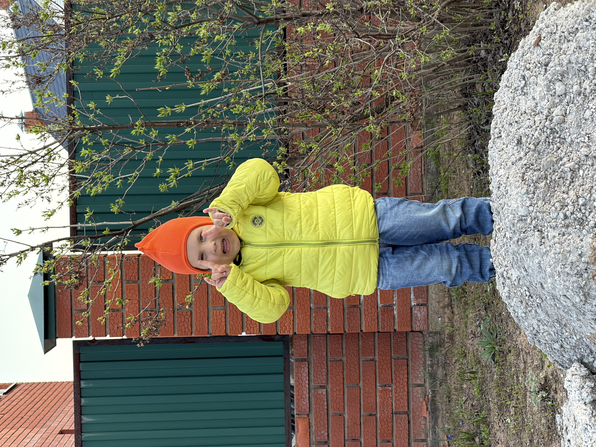 A young child in a yellow jacket and orange hat stands sideways in an outdoor setting. The child is making a gesture with their hands near their face. A green and brown fence is visible in the background, along with a brick wall and a large pile of gray aggregate. The location is Chaikovsky, Russia.