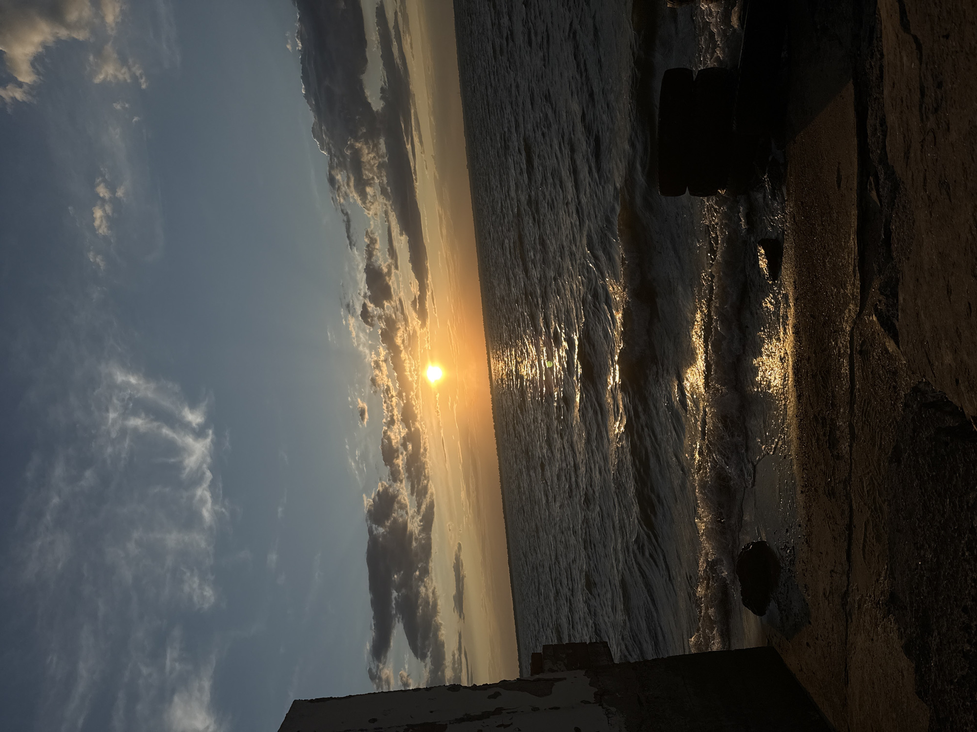 This is a photograph of a sunset over the Black Sea in Zatoka, Ukraine. The sun is low on the horizon, casting a golden light across the water and illuminating the clouds. The waves are small and gentle, lapping at the shore. In the foreground, there is a concrete structure on the left and two tires stacked on the right. The overall mood is peaceful and serene.