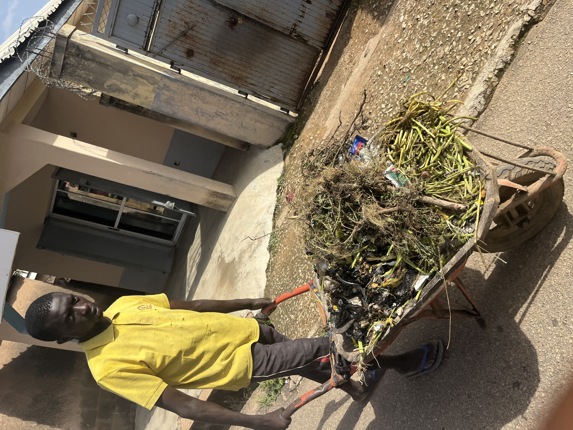 A young Black male wearing a yellow polo shirt and dark shorts is pushing a wheelbarrow filled with debris. The wheelbarrow contains a mixture of plant matter, plastic bags, and other waste. The man is wearing blue flip-flops. The background shows a building with a beige facade and a metal gate with barbed wire on top, consistent with a location in Jos, Nigeria. The ground is paved and a shadow of the man and wheelbarrow is cast on it. The image appears to be taken in an outdoor urban or semi-urban setting.