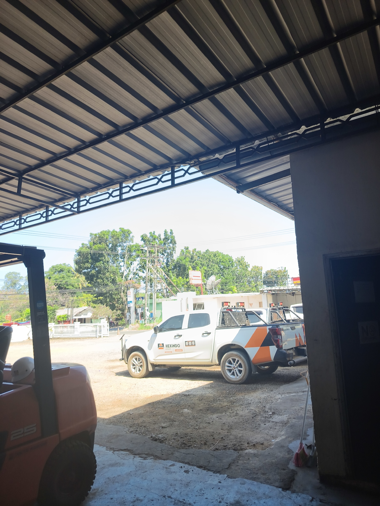 The image depicts an outdoor scene at a workshop or industrial area, possibly in Pangkalpinang, Indonesia. In the foreground, a red forklift is parked, its front visible. Behind it, two white pickup trucks with orange and black decals are parked. The trucks have equipment racks on their beds and some sort of signage on the doors. One truck has the logo "HEXINDO" visible. The ground is dirt and gravel. Overhead, a corrugated metal roof supported by a metal frame covers the area, creating shadows. In the background, there are trees, utility poles, and some buildings. The sky is clear and bright. A broom is leaning against a wall on the right side of the image. No people are clearly visible in the scene.