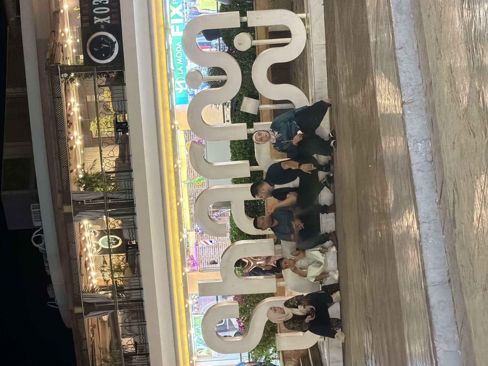 The image captures an outdoor scene at night in Sharm El Sheikh, Egypt, featuring a large, illuminated, white sculptural sign with Arabic lettering. Several individuals are seated on and around the sculpture. On the left, a woman wearing a hijab and a patterned abaya sits with her legs tucked under her. Next to her is a young girl in a light-colored dress. To their right, two men are seated, one in a dark shirt and the other in a black t-shirt and dark pants. Further to the right, a woman wearing a teal hijab and a matching outfit is seated. In the background, a multi-story building is visible, illuminated by strings of lights. Above the large sculpture, a bright sign displays the words "LA MODA FIX." The ground surface appears to be wet, reflecting the ambient lighting.
