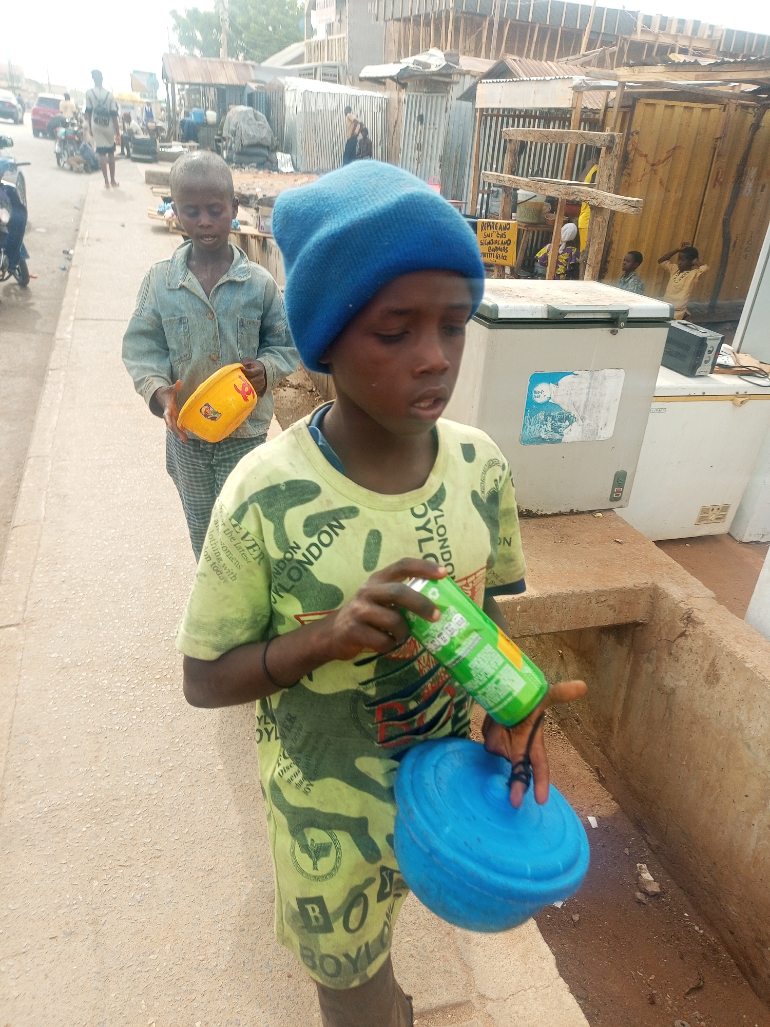 The image captures a daytime street scene, likely in an urban or semi-urban area such as Tempure, Nigeria, under an overcast or hazy sky. Two young boys are the primary subjects, walking along a paved path next to a road.
The boy in the foreground, on the right, wears a blue knit beanie and a light green, camouflage-patterned t-shirt. The t-shirt features text such as "YLONDON," "BOYLO," and phrases like "EVER the latest womens Nothing with on today." He holds a green beverage can in his right hand and a blue plastic bowl or lid in his left. His expression is neutral.
Behind him, slightly to the left, another boy with a close-cropped haircut wears a light blue denim jacket over striped trousers. He carries a yellow plastic bowl, which has a red symbol resembling a flower or star on its side. He also has a neutral expression.
The background reveals a bustling street environment. Motorbikes are visible on the road, and other people, including adults and children, can be seen. The roadside is lined with simple structures, some made of corrugated iron sheets and wood, indicative of market stalls or small shops. Two large white chest freezers are positioned near a concrete drain, one bearing a "Big-F" label and a blue sticker. A yellow shipping container also forms part of the structures. A visible sign, attached to a wooden frame, reads: "REPIRE AND SALE CLAS SILNOIRS AND BORNERS 070117 6362," advertising repair and sales services. The overall scene depicts daily life and commerce in a busy, roadside setting.