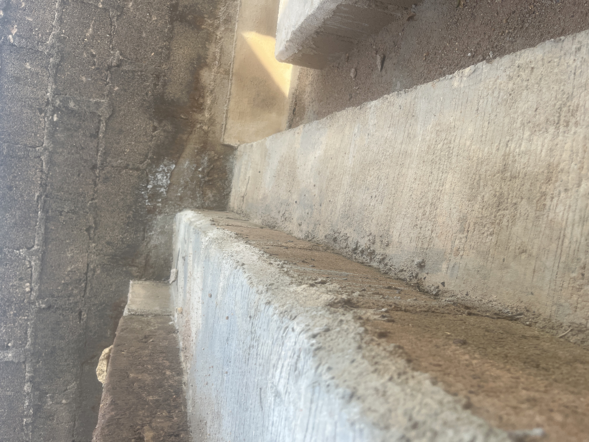 The image shows a close-up view of a series of concrete steps leading upwards. The steps are weathered and stained, showing signs of age and exposure. Soil and debris are visible on the steps and adjacent surfaces. The wall to the left is made of a rough, dark-colored material, possibly concrete or stone, with noticeable texture and variations in color. The steps ascend away from the viewer, creating a forced perspective. The location, Barakin Akawo, Nigeria, is provided externally, suggesting this structure is situated in that city.