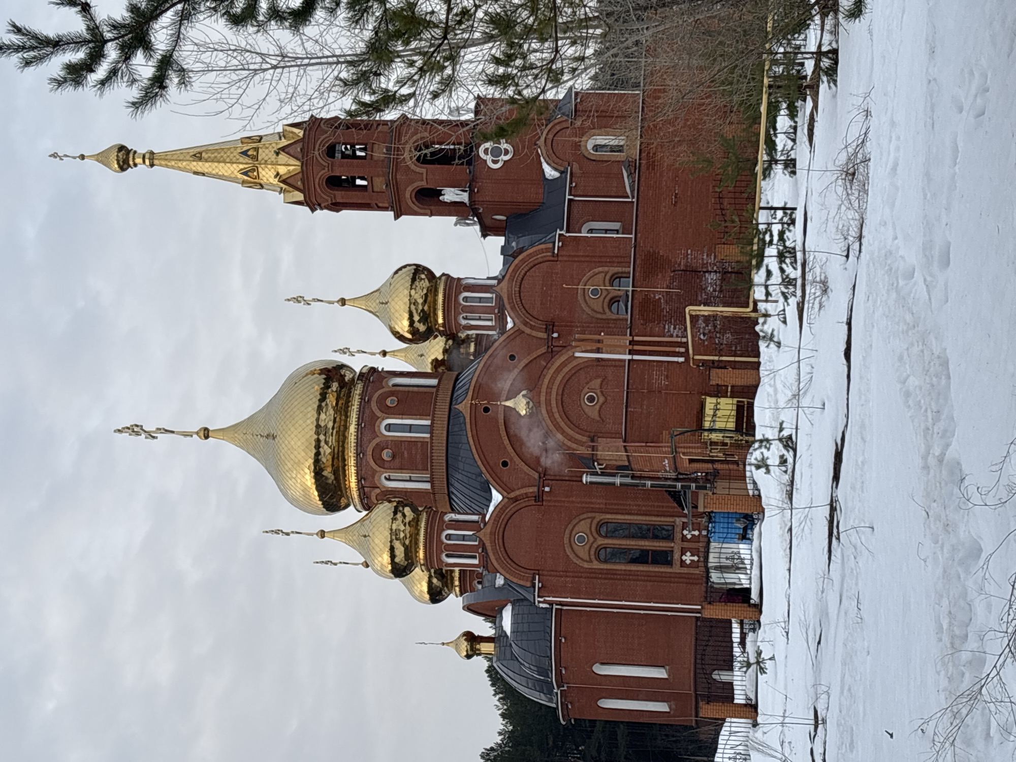 The image depicts the Assumption Cathedral (Uspensky Sobor) located in Chaikovsky, Perm Krai, Russia, observed during a winter or early spring season. The large, multi-domed Orthodox church is constructed primarily of red brick. Seven prominent golden, onion-shaped domes, each topped with an Orthodox cross, rise above the main structure, with smaller dark gray roofs connecting them. A tall, slender bell tower stands on the right, capped with a golden spire and cross, and features multiple arched openings containing bells. A white, angelic statue is visible on a ledge of the bell tower. White steam or smoke is emanating from a pipe located on the lower-mid section of the church's right facade. The foreground consists of a thick, even layer of snow, displaying faint footprints and scattered, sparse vegetation including small coniferous trees and bare deciduous branches. A dark metal fence delineates the church property, behind which yellow utility pipes are affixed to the brick wall. A blue barrel-like object and a yellow cuboid container are visible behind the fence near the church's base. A dense coniferous forest forms the background on the left, while bare deciduous trees are present on the right. The sky is uniformly overcast and light gray. No individuals are present in the scene, and no specific actions or interactions are depicted.