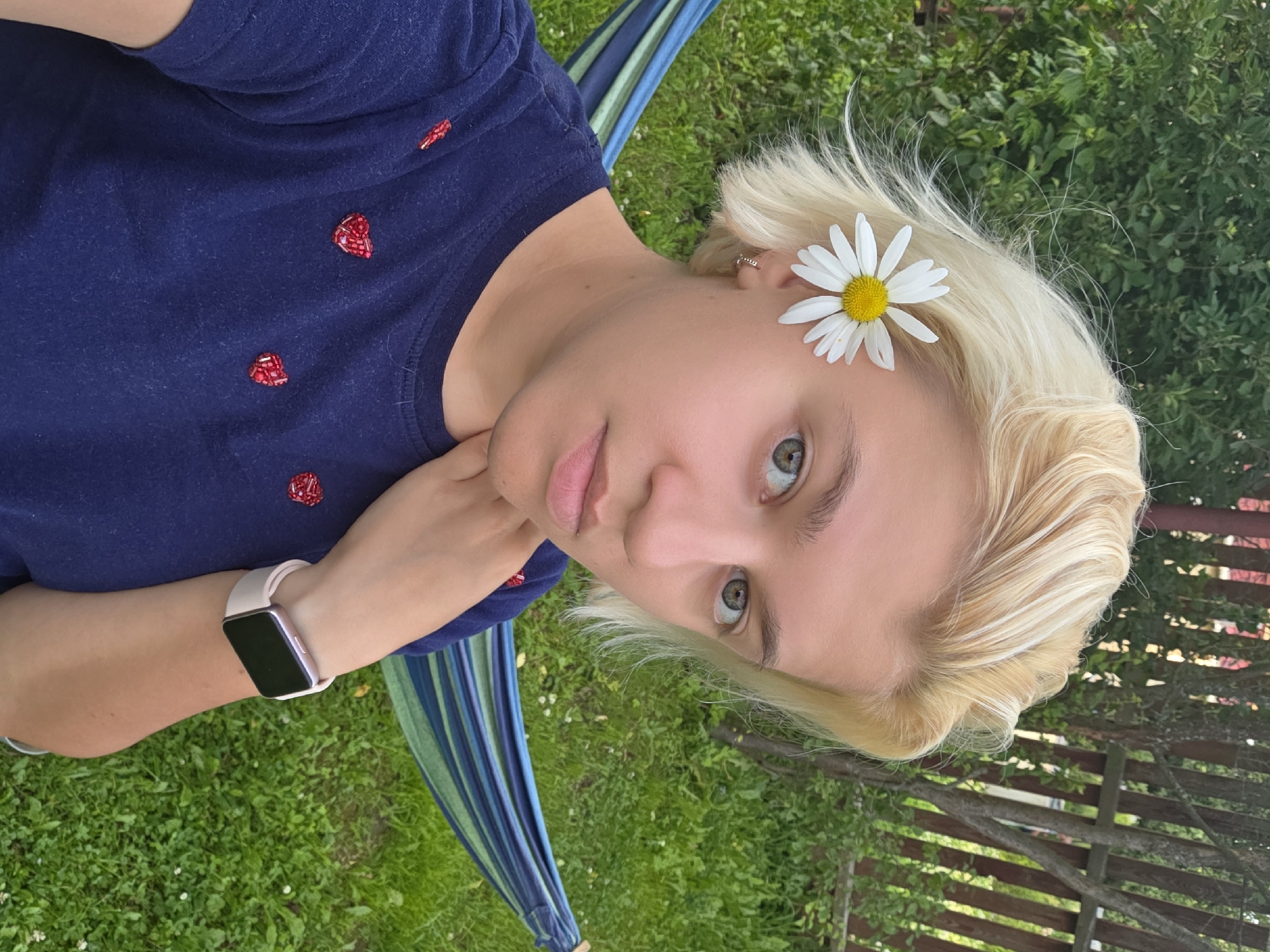 A young woman with short, blonde hair is captured in a selfie outdoors. A white daisy with a yellow center is tucked behind her left ear. She is wearing a navy blue t-shirt adorned with small, red, heart-shaped embellishments. On her left wrist, she wears a light-colored smartwatch. The woman is leaning her right hand on her chin, looking towards the camera with a neutral expression. Behind her, a hammock with blue and green stripes is suspended between two points, set against a backdrop of lush green grass and trees, with a wooden fence visible in the upper right quadrant. This scene appears to be set in Frolovo, Russia.
