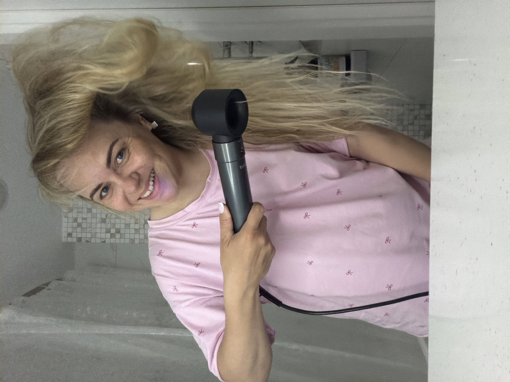 A woman is holding a Dyson Supersonic hair dryer in a bathroom setting. She is wearing a light pink shirt with a small bow pattern and has long blonde hair. She is smiling, and her hair is flipped upwards. The bathroom has white walls and a tiled area. The image has been rotated, so the scene appears sideways. The location in the city of Votkinsk, Russia cannot be determined from the image.