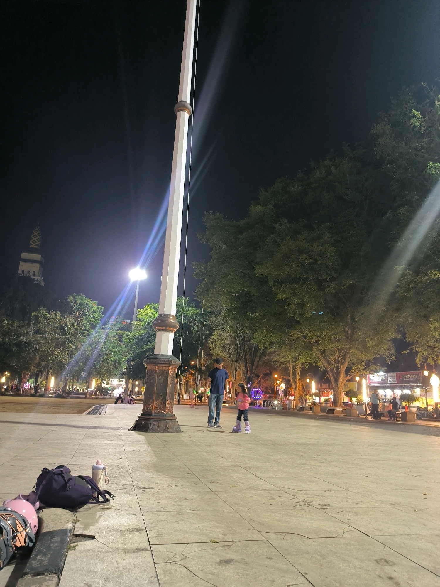 The image depicts a nighttime scene in Salatiga, Indonesia. In the foreground, a large flagpole stands centrally, its white shaft contrasting with the dark sky. To the left, a dark backpack and a water bottle rest on the paved ground, alongside two helmets, one pink and one grey and black. A man wearing a blue t-shirt and jeans stands to the right of the flagpole, and a young girl in a pink jacket and rollerblades is positioned in front of him, seemingly learning to skate or playing.
The background reveals a park-like setting with numerous trees, some illuminated by streetlights. The warm glow of lights from shops and food stalls is visible in the distance, creating a lively atmosphere. A distinct tower with an ornate, patterned cap is visible in the far left background, a prominent landmark. People are scattered throughout the background, seated at tables or strolling, suggesting a public gathering space. The overall environment is an urban public area at night, likely a town square or park, buzzing with evening activity.