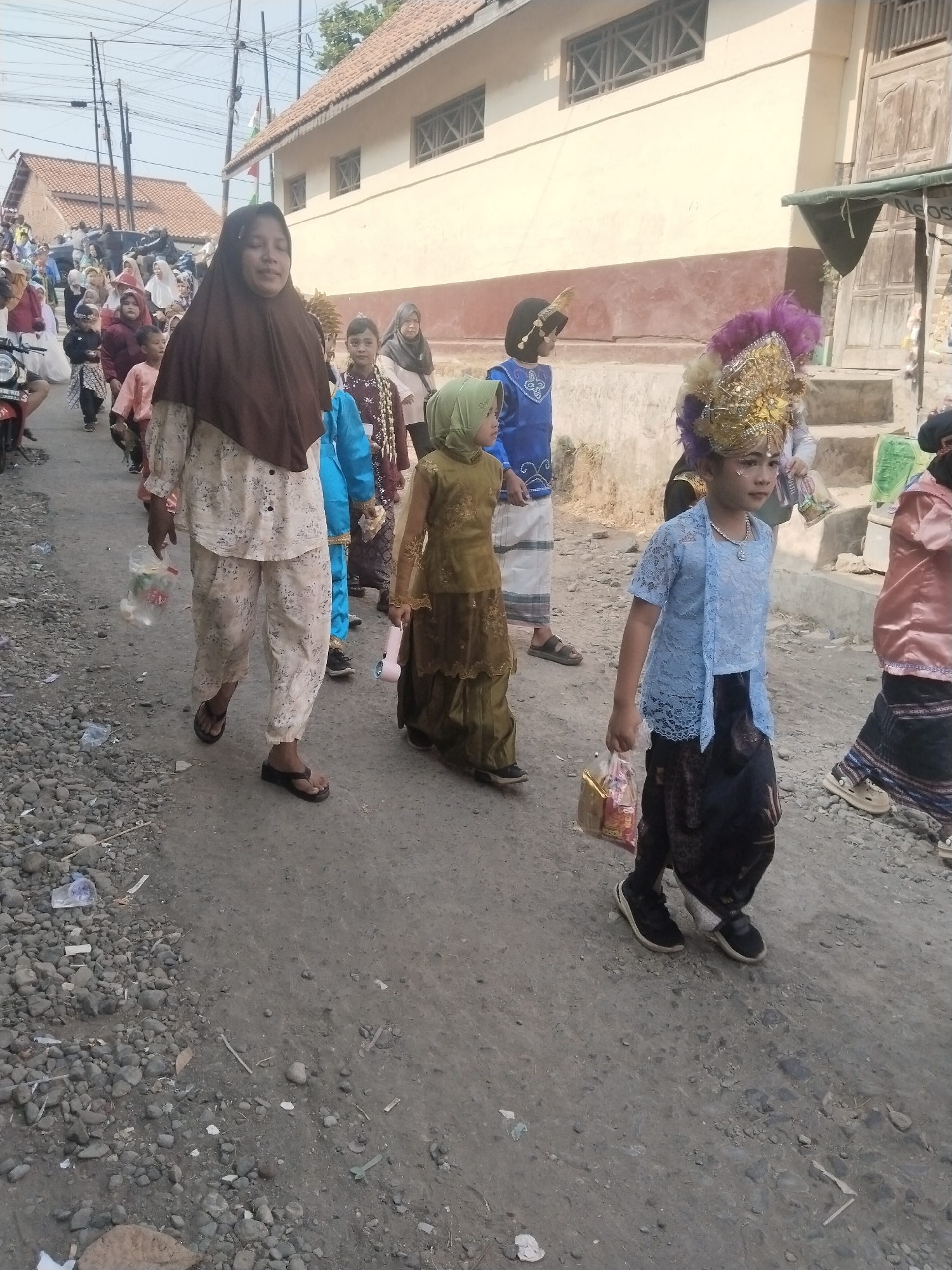In the city of Jembayat, Indonesia, a procession of people, predominantly children, walks down a dusty, graveled street. The event appears to be a cultural or religious celebration, as many of the participants are dressed in traditional attire, including intricately decorated headwear and vibrant clothing.
In the foreground, a young girl wearing a light blue traditional top and a dark patterned sarong walks with confidence, her face adorned with makeup. She carries a small golden gift bag. To her right, another child, also in traditional dress and an elaborate golden headpiece with purple feathers, is partially visible.
Behind them, a woman in a brown hijab and a patterned outfit walks, holding a clear plastic bag with items inside. Several other children in various traditional outfits are seen participating in the procession, some carrying small gifts. The background features simple, single-story buildings with tiled roofs, typical of the local architecture. Overhead, power lines crisscross the sky, indicating the presence of modern infrastructure in the rural setting. The overall scene depicts a lively community event taking place in a modest urban environment.
