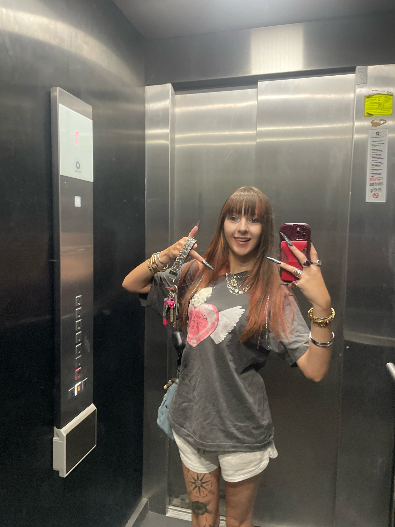 The image depicts a young woman taking a selfie in an elevator. She is wearing a dark grey t-shirt with a graphic print of a winged heart, white shorts, and several pieces of jewelry including necklaces, bracelets, and rings. She has long hair with streaks of red and bangs, and long artificial nails. She is holding a smartphone in her right hand and making a hand gesture with her left. The elevator has stainless steel walls and a dark panel with buttons and a digital display on the left. There are also some stickers on the elevator door. The scene appears to be located in an urban setting.