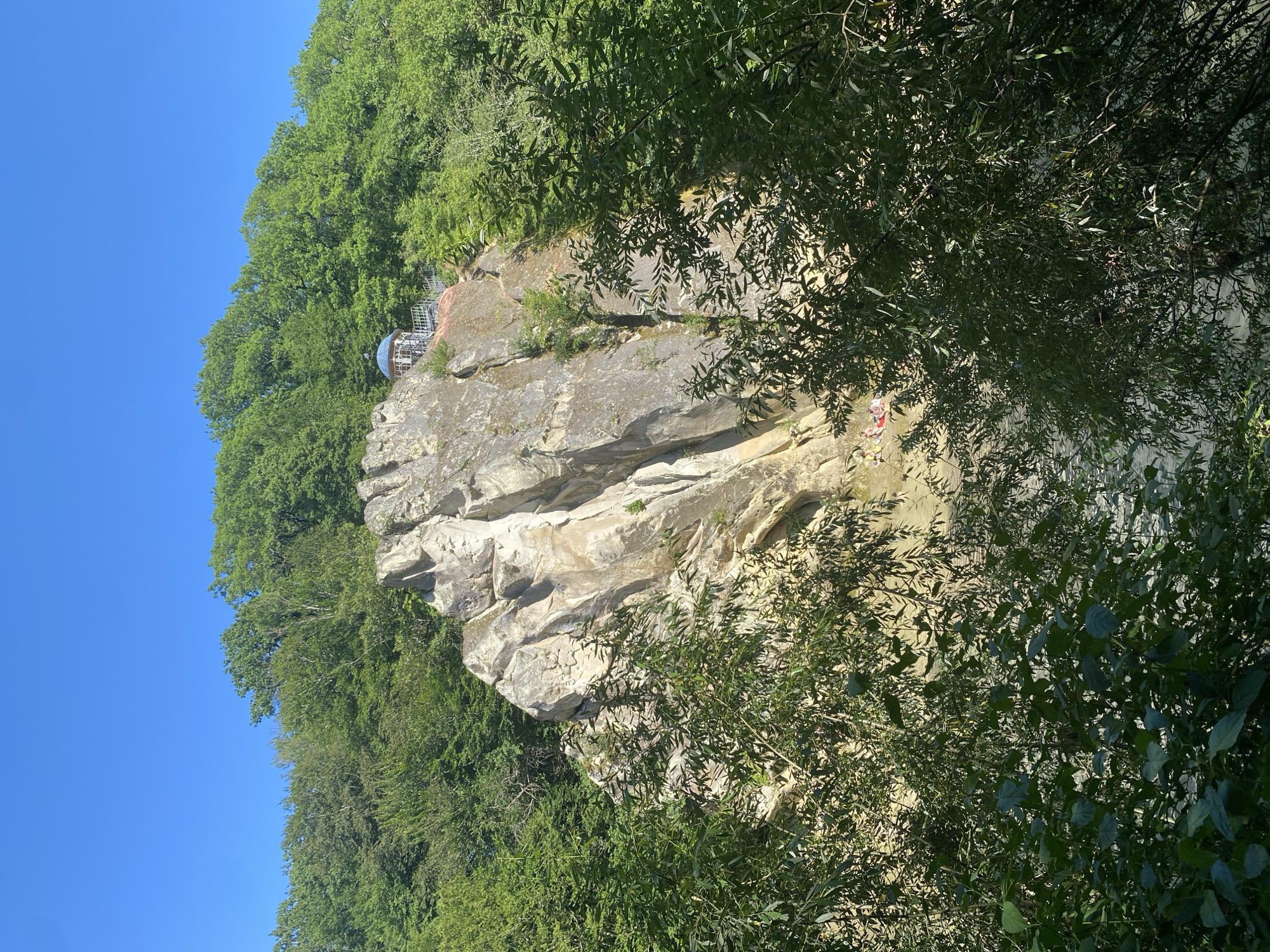 The image depicts a natural landscape with a prominent rock formation. A small, ornate gazebo or structure with a blue domed roof is situated atop the rock formation, accessible by a railing and steps. Lush green trees and foliage surround the rock and extend to the top of the frame, contrasting with the clear blue sky. At the base of the rock formation, near a body of water, several individuals are visible, some appearing to be sunbathing or relaxing on the shore. The scene suggests a recreational area within a natural environment. This location is known as the "Divny Stone" in Goryachiy Klyuch, Russia, a popular natural landmark.