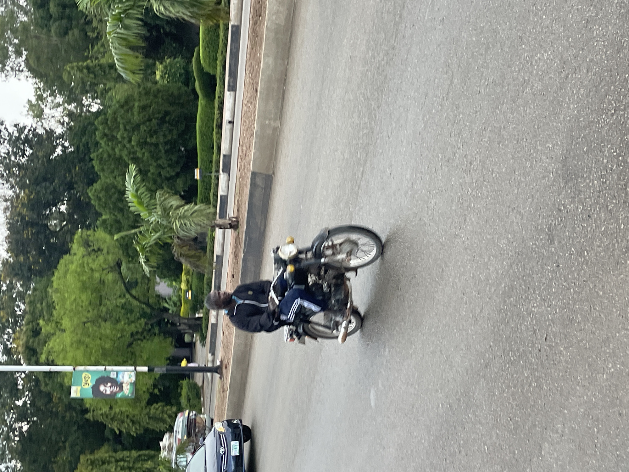 A medium shot captures a man riding a motorcycle on a paved road, likely in Jos, Nigeria, given the context of previous images. The rider, a Black male with a dark complexion, is wearing a black jacket with white stripes on the sleeves and dark pants with white stripes down the legs. He is seated on a black motorcycle. In the background, lush green trees form a dense backdrop, suggesting a well-maintained urban park or roadside. A tall lamppost with a banner featuring a smiling woman and the word "YES!" is visible to the left. A dark blue car is also present on the road to the left of the motorcycle. The road surface appears slightly wet, indicating recent rain or damp conditions. The overall atmosphere is that of a typical daytime scene in a Nigerian city.