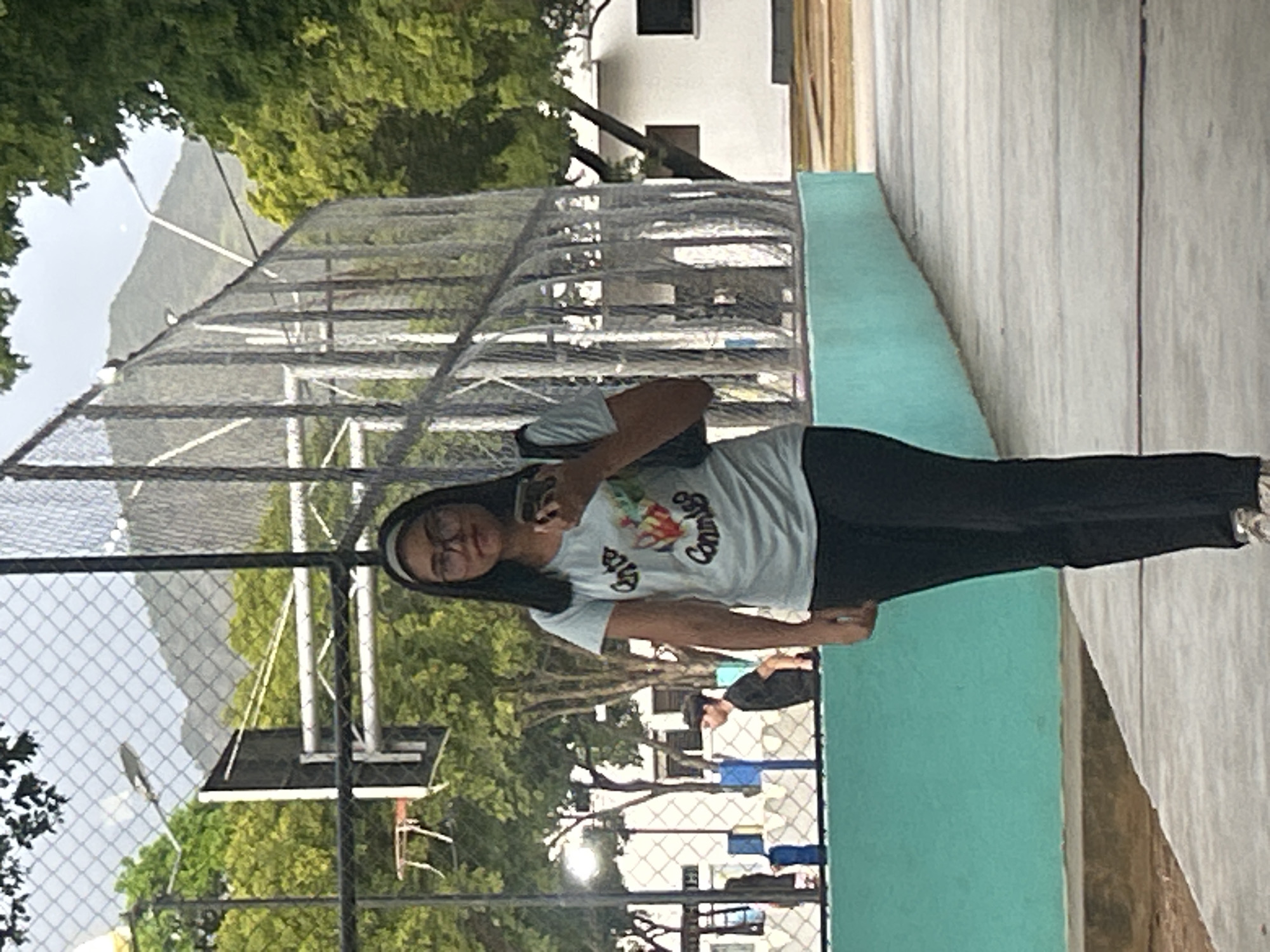 A young woman wearing glasses, a light blue t-shirt with graphics, and black pants stands on a paved walkway. She holds a smartphone in her right hand and has a black bag slung over her left shoulder. The background features a chain-link fence, trees, a basketball hoop, and buildings. In the distance, a mountain is visible. The scene appears to be an outdoor recreational area, possibly a school or park, in Santa Ana, Venezuela.