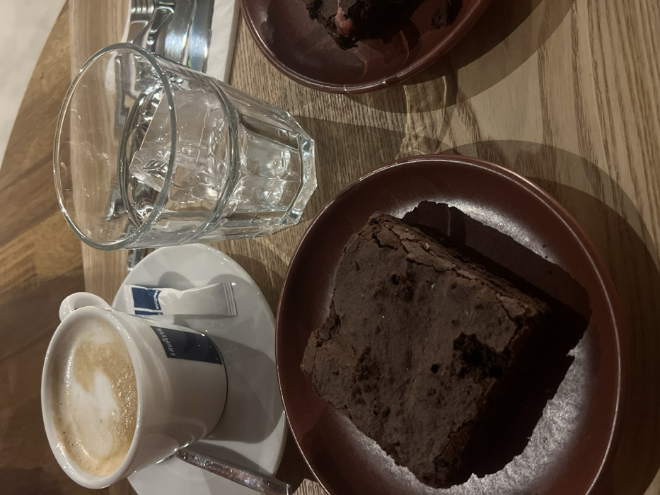 The image displays a tabletop scene within an establishment located in Lordelo do Ouro e Massarelos, Portugal. The surface is a light-colored wooden table. A white ceramic cup bearing a blue "Lavazza" logo contains a cappuccino with distinct foam art and rests on a white saucer. A silver spoon and a "Lavazza" branded sugar packet are positioned on the saucer. Adjacent to this, a clear, faceted glass tumbler holds water and several ice cubes. In the lower left, a dark chocolate brownie with a cracked surface is presented on a dark red circular plate. A second dark red plate, partially visible in the lower right, contains a partially consumed chocolate brownie, which exhibits a reddish inclusion. Objects in the background, including cutlery and a white napkin, are out of focus. No individuals are present in the frame.