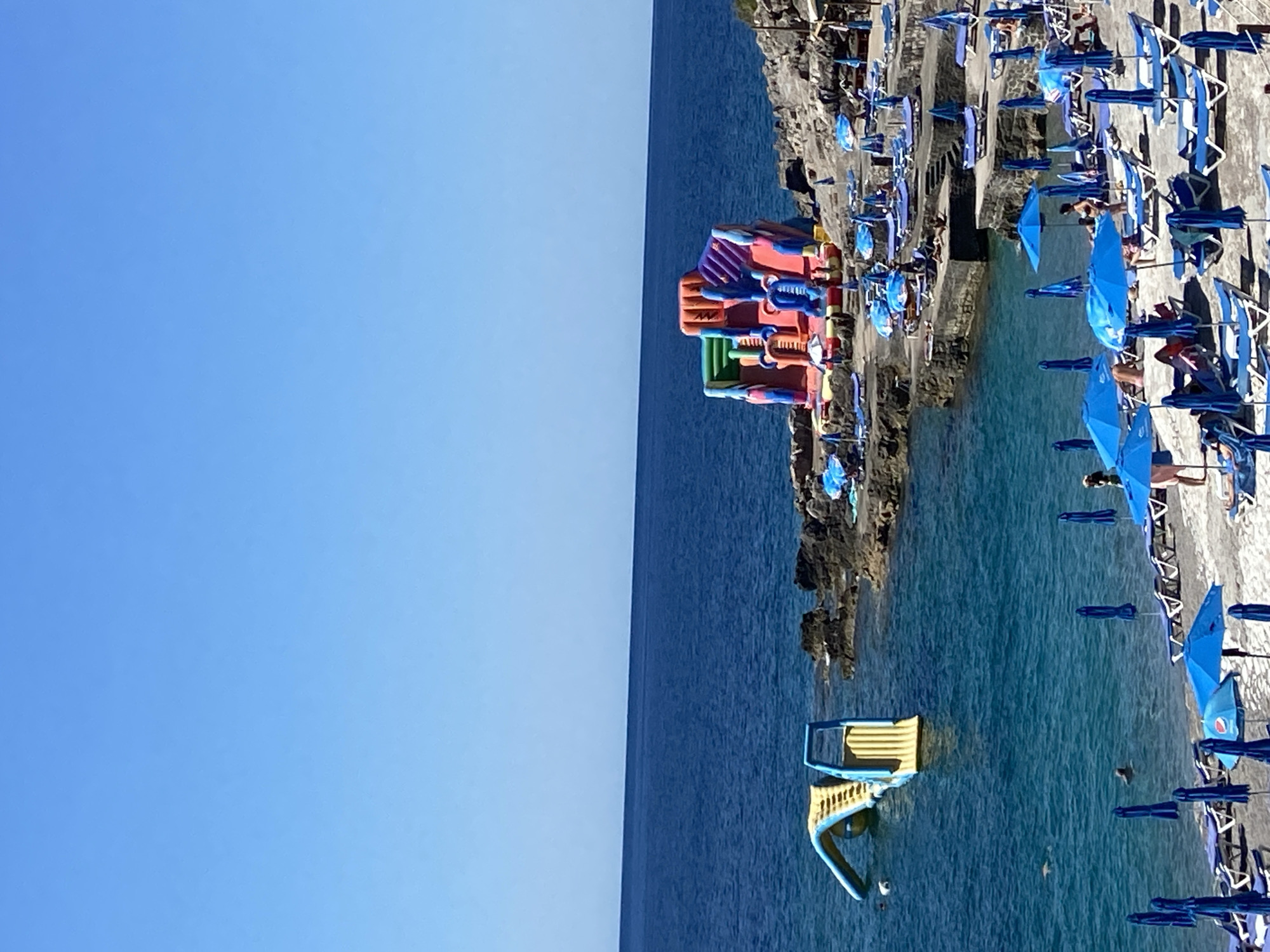 The image depicts a sunny day at a beach with clear blue waters and a bright blue sky. In the foreground, a beach lined with numerous blue lounge chairs and umbrellas is visible. The chairs are mostly empty, with a few people scattered among them, some sunbathing and others sitting. The water is calm, with several people swimming near the shore and a yellow inflatable water slide extending from the beach into the sea on the left side of the image. Further out in the water, a large, colorful inflatable playground structure with slides is anchored. The beach is situated along a rocky coastline, with stone steps leading up from the water to the beach area. In the background, the dark blue sea stretches to the horizon, meeting the clear blue sky. The scene suggests a recreational area in Prijevor, Montenegro, designed for beachgoers and water activities.