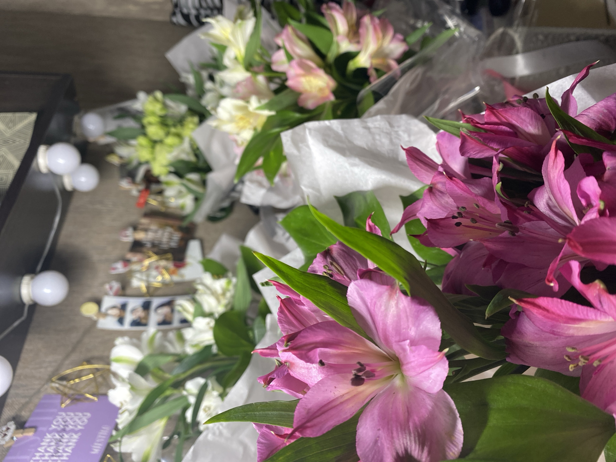 The image shows several bouquets of flowers. The bouquets include pink, white, and green flowers, wrapped in white paper and clear plastic. A vanity mirror with light bulbs is visible on the left side. A purple card with "THANK YOU" written on it and small photo strips are placed near the mirror. The location appears to be indoors. There is no information in the image to determine the specific location in the city of Chaikovsky, Russia.