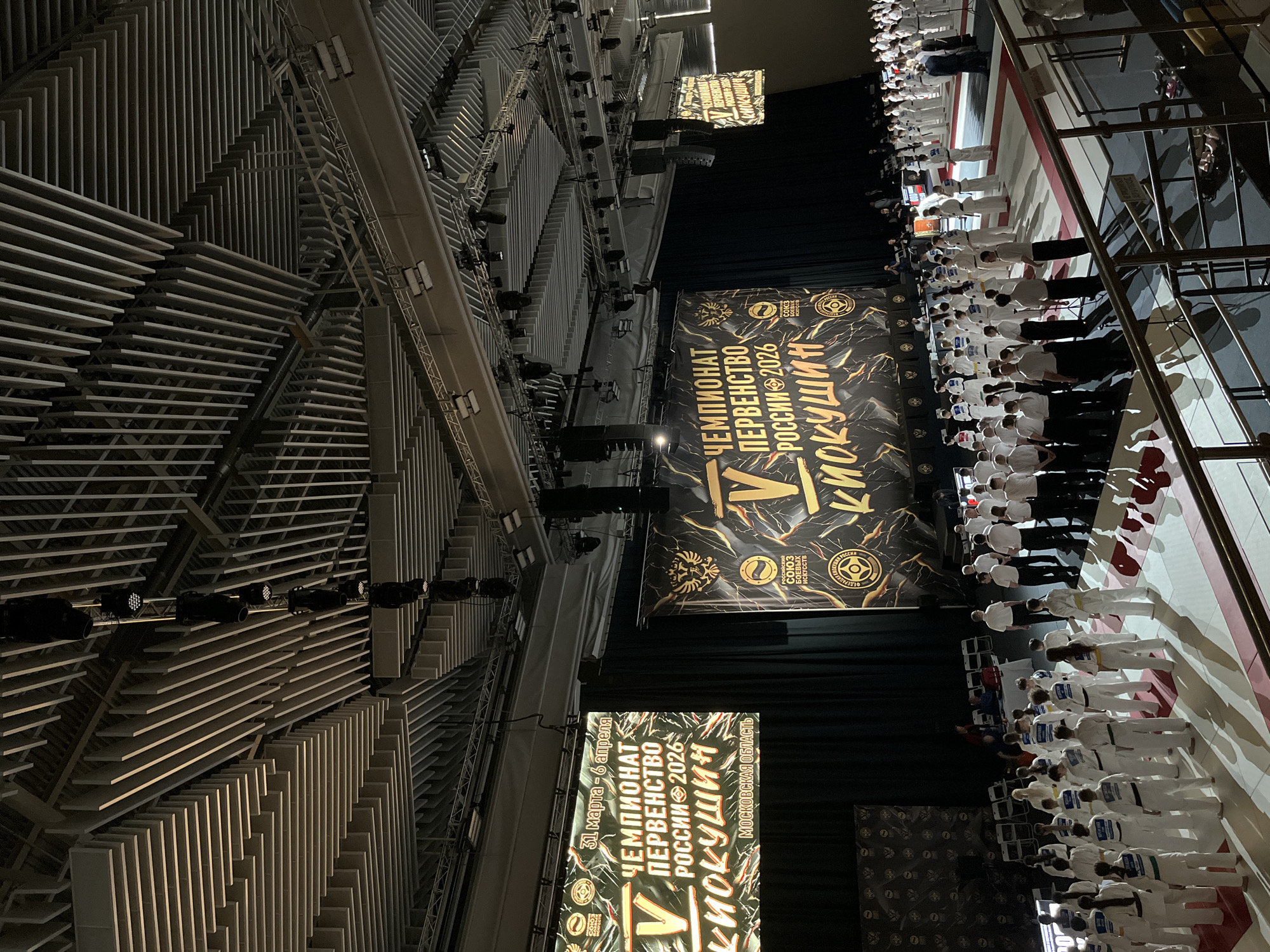 The image depicts an indoor martial arts event, likely a Kyokushin Karate tournament, in Zhavoronki, Russia. A large banner hangs in the background, displaying the event's title "Чемпионат Первенство России 2026 Киокушин" (Championship of the Russian Federation 2026 Kyokushin) and the dates "31 марта - 6 апреля" (March 31 - April 6). Below it, athletes wearing white uniforms stand in rows. Above the banner, a rigging system with lights is visible. The building has a modern architectural style with geometric designs on the walls and ceiling.