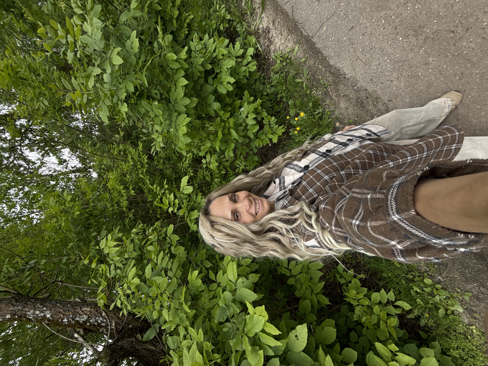 A woman with blonde hair is outdoors, possibly in Rusanovo, Russia. She is smiling and wearing a brown and white plaid shirt. She is partially surrounded by green leafy bushes and a tree trunk. A paved path is visible along the edge of the greenery. One of her feet, wearing a tan shoe, is visible on the path.