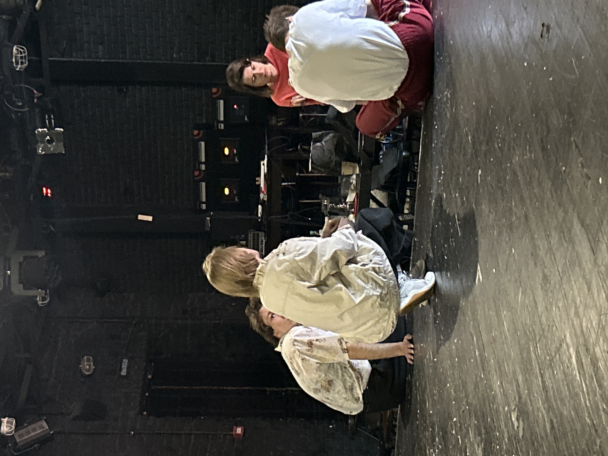 In an indoor setting with dark, brick walls and a worn wooden stage, four individuals are seated in a circle. The person furthest to the left wears a light-colored shirt with a pattern. Next to them, facing away from the camera, is someone in a beige jacket. To their right, a woman in an orange top looks down. The person on the far right, facing away, is wearing a white t-shirt and red pants. A table with various items, including a glass and bags, is visible behind the seated group. The environment suggests a theater or rehearsal space.