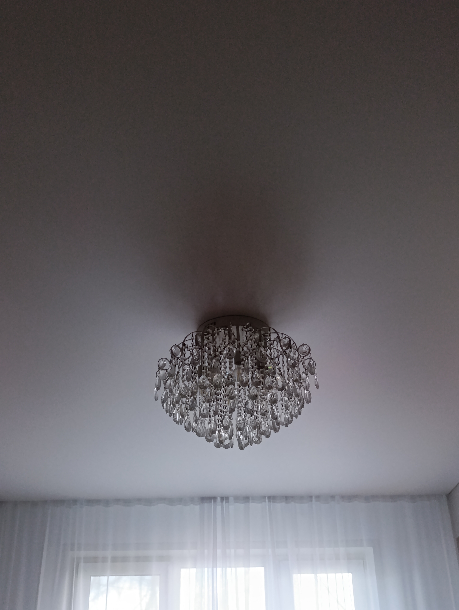 A low-angle interior shot showcases a crystal chandelier affixed to a white ceiling. Light diffuses through sheer white curtains, partially obscuring a window below. The location is indicated as Krasnoyarsk, Russia. No people or actions are visible.