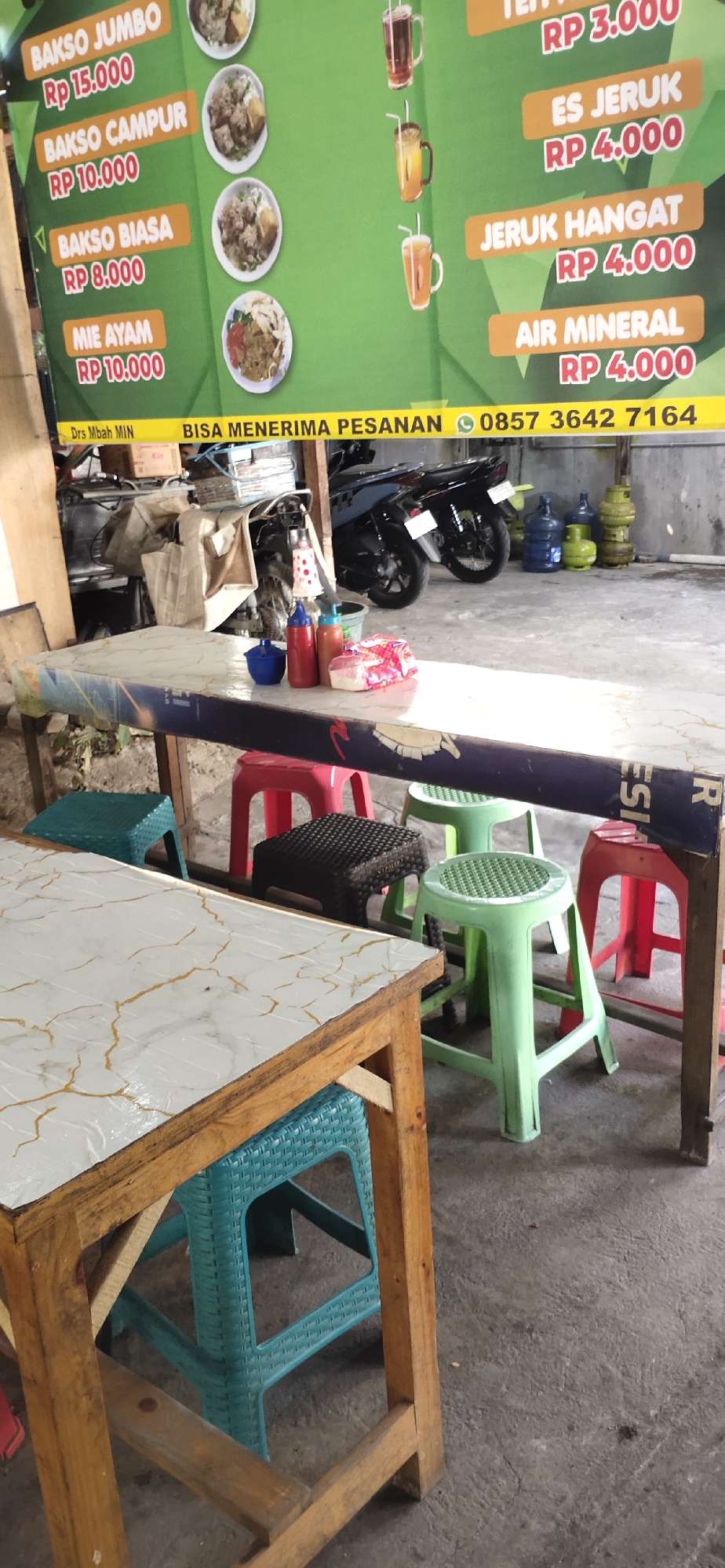The image captures a close-up view of a table and chairs set up outdoors, likely in a food stall or small eatery. The table, covered with a white marble-patterned cloth, is positioned in the foreground. Several plastic stools in various colors (teal, red, green, and black) are arranged around the table. On the table, condiments such as chili sauce and ketchup are visible, along with a package of tissues.
In the background, a large green banner displays a menu with food and drink items, including "Bakso Jumbo," "Bakso Campur," "Bakso Biasa," "Mie Ayam," "Es Jeruk," "Jeruk Hangat," and "Air Mineral," along with their prices. To the right of the banner, several large water jugs are stacked. A motorcycle is parked behind the table.
The overall environment appears to be a casual, outdoor setting with a concrete floor and some rustic wooden structures supporting the table. The presence of the menu and the seating arrangement suggests this is a place where customers can dine.
Based on visual cues such as the Indonesian language on the menu and the general aesthetic of the establishment, it is plausible that this scene is located in the city of Mojokerto, Indonesia. The banner also includes the text "BISA MENERIMA PESANAN 0857 3642 7164", which translates to "CAN RECEIVE ORDERS", indicating a business that takes orders. The mention of "Drs. Mbah MIN" likely refers to the proprietor or name of the establishment.