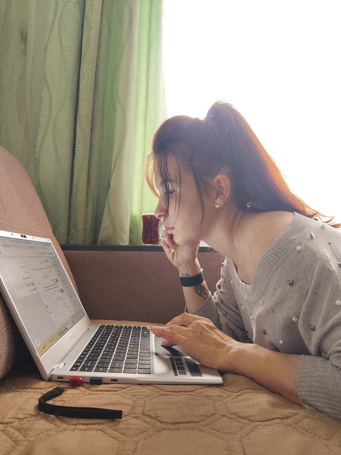 This image shows a young woman intently working on a laptop indoors, likely at home in Фоки, Russia. She is seen from the right side, reclining on a brown, quilted surface, which appears to be a couch or bed.
The woman has long, dark hair styled in a high ponytail, which is brightly lit from behind by a strong light source. She is wearing a light grey, long-sleeved sweater embellished with small white pearls or beads. A gold-colored earring is visible in her right ear, and a dark-colored wristband or fitness tracker is on her left wrist, beneath which a tattoo can be seen. Her fingernails are manicured with a light polish.
She is deeply engrossed in the silver-colored laptop positioned directly in front of her. Her left hand supports her chin, indicating focus, while her right hand rests on the laptop's touchpad or keyboard, suggesting active use. A red USB drive is plugged into the left side of the laptop, and a black wrist strap lies on the brown surface next to it. The laptop screen displays what appears to be a web interface or application with various text fields and elements, though the specific content is unreadable. A partial "ACER" logo is visible on the laptop's hinge.
In the background, a small glass containing a dark red liquid, possibly tea, is placed on the furniture behind her. Beyond that, light green curtains with a prominent swirling pattern hang in front of a very bright window, indicating that it is daytime. The strong backlighting from the window creates a significant contrast and a luminous effect around the woman's hair. The overall scene suggests a comfortable, focused work or study session.